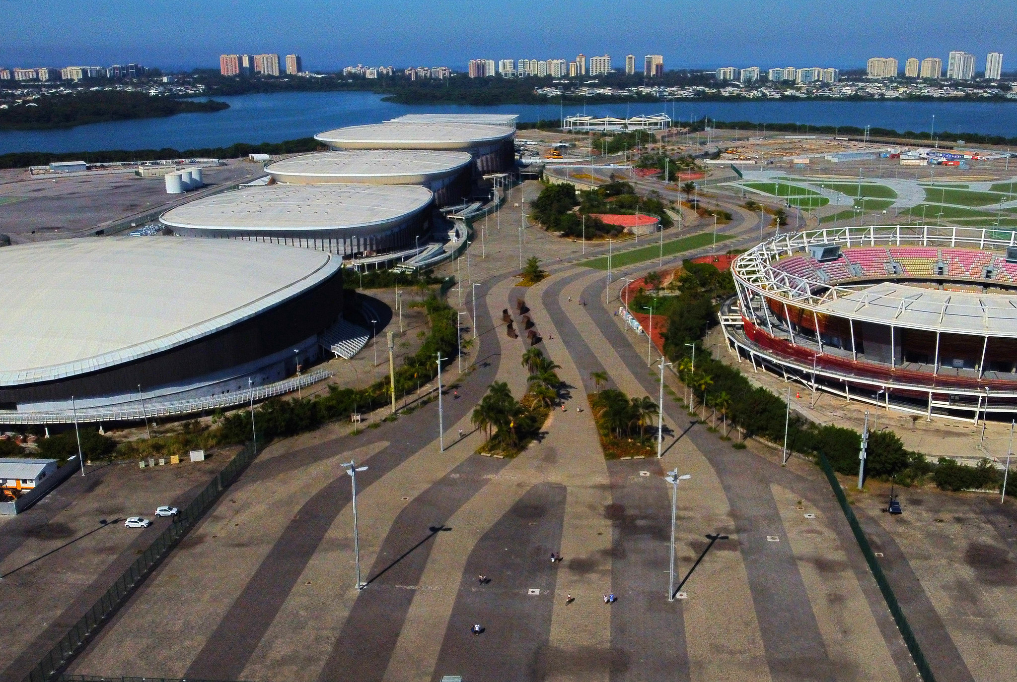 Uma vista aérea do Parque Olímpico na Barra Olímpica, Rio de Janeiro, mostrando grandes edifícios de teto branco, um estádio circular, estradas e áreas verdes, com um lago e a cidade ao fundo.