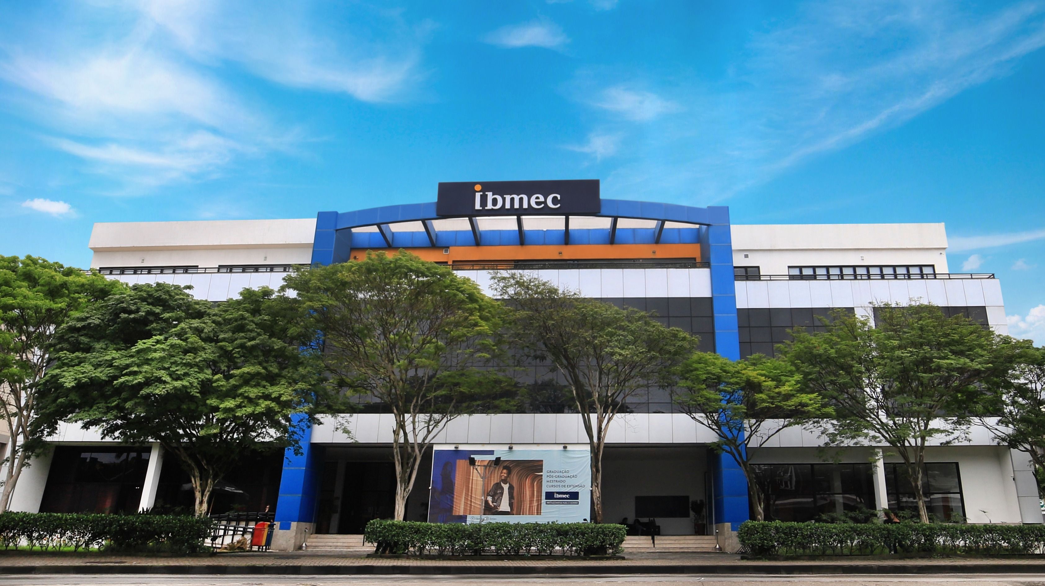 A fachada de um prédio moderno com o nome "IBMEC" em destaque, cercado por árvores e arbustos, sob um céu azul claro.