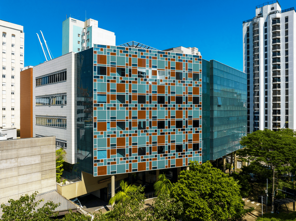 Vista do edifício Hospital Albert Einstein em Perdizes, com uma fachada de vidro e painéis coloridos em tons de azul e marrom, destacando-se entre outros prédios altos. Árvores verdes e o céu azul completam a cena urbana.