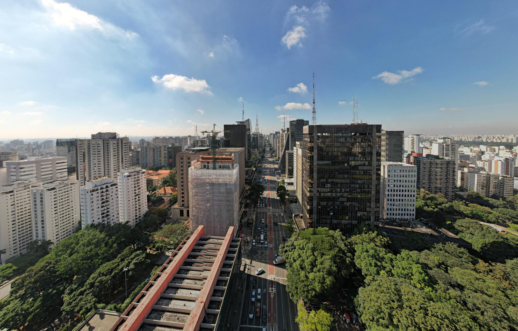 Vista panorâmica da Avenida Paulista, com arranha-céus e tráfego intenso em uma rua movimentada. Árvores verdes margeiam a avenida, e o céu azul com nuvens esparsas completa a paisagem urbana.