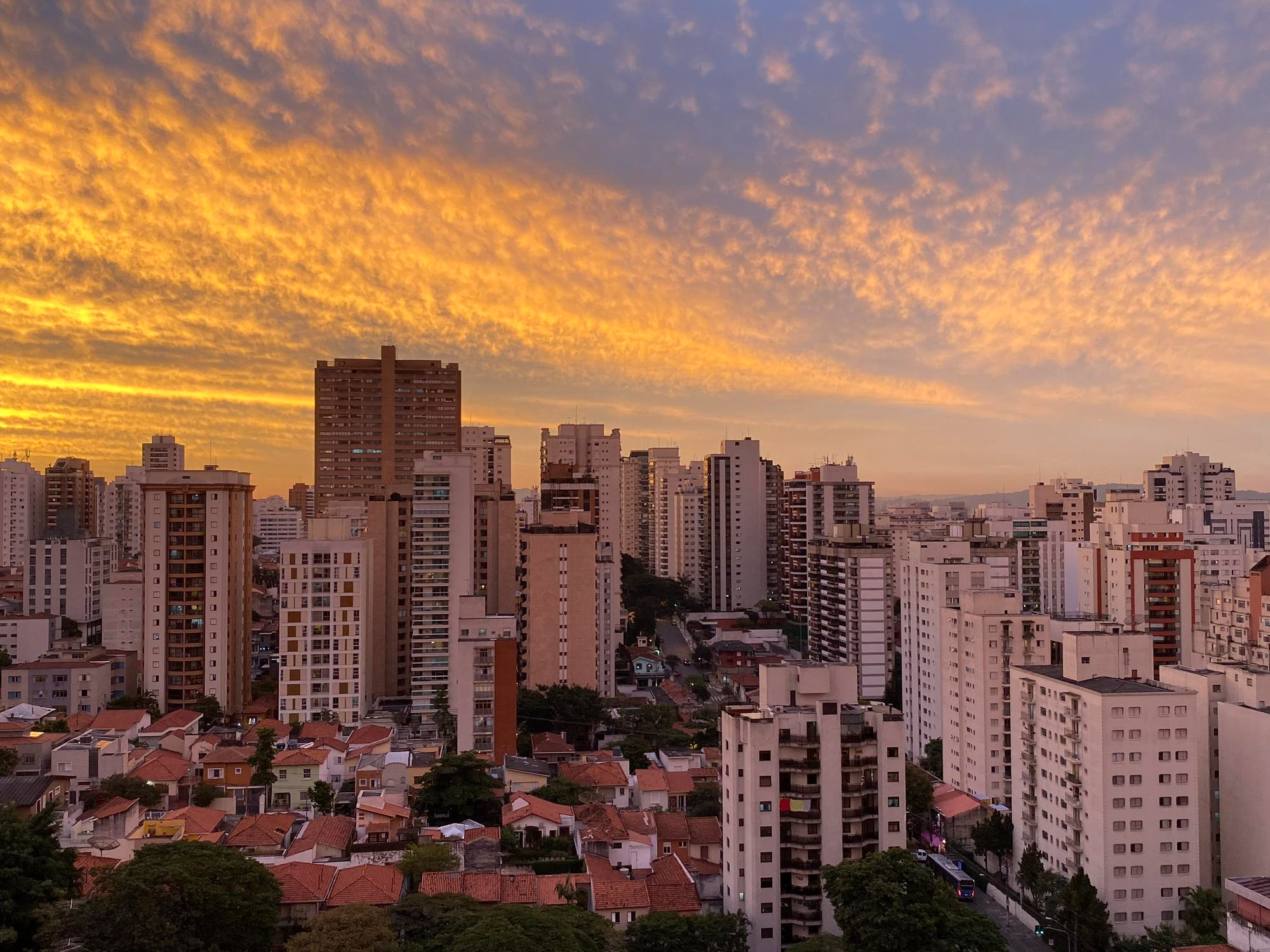 Vista aérea de uma cidade ao pôr do sol, com o céu em tons de laranja e dourado sobre uma paisagem urbana densa de edifícios altos.