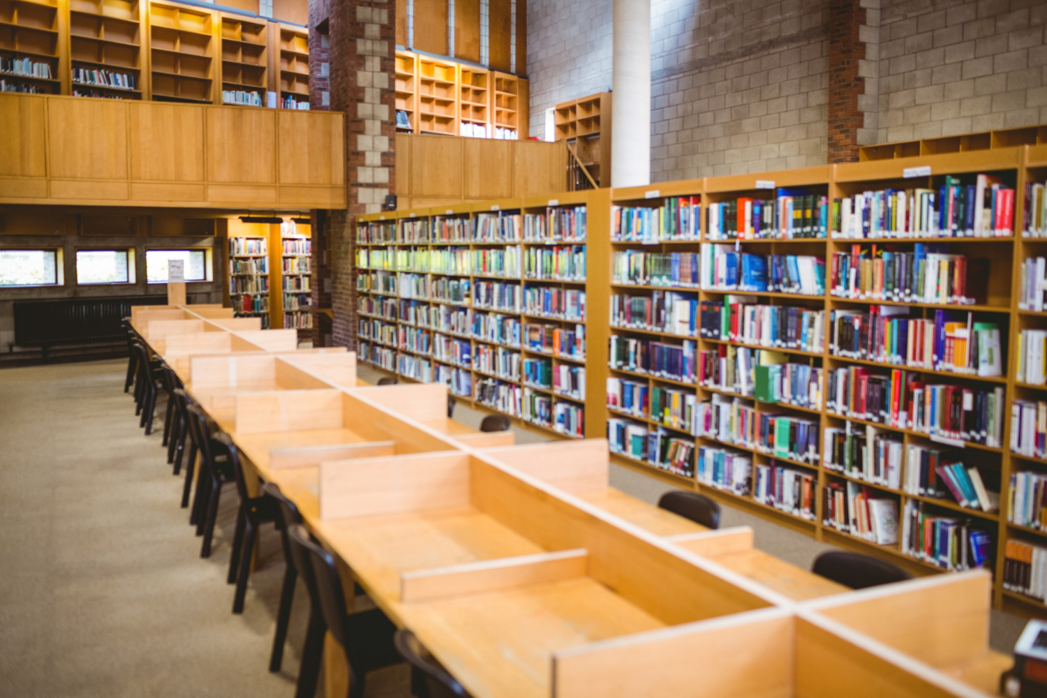 Uma biblioteca espaçosa com longas mesas de estudo de madeira e cadeiras pretas, ladeadas por estantes repletas de livros. A arquitetura apresenta detalhes em tijolo e madeira, com uma área superior visível.