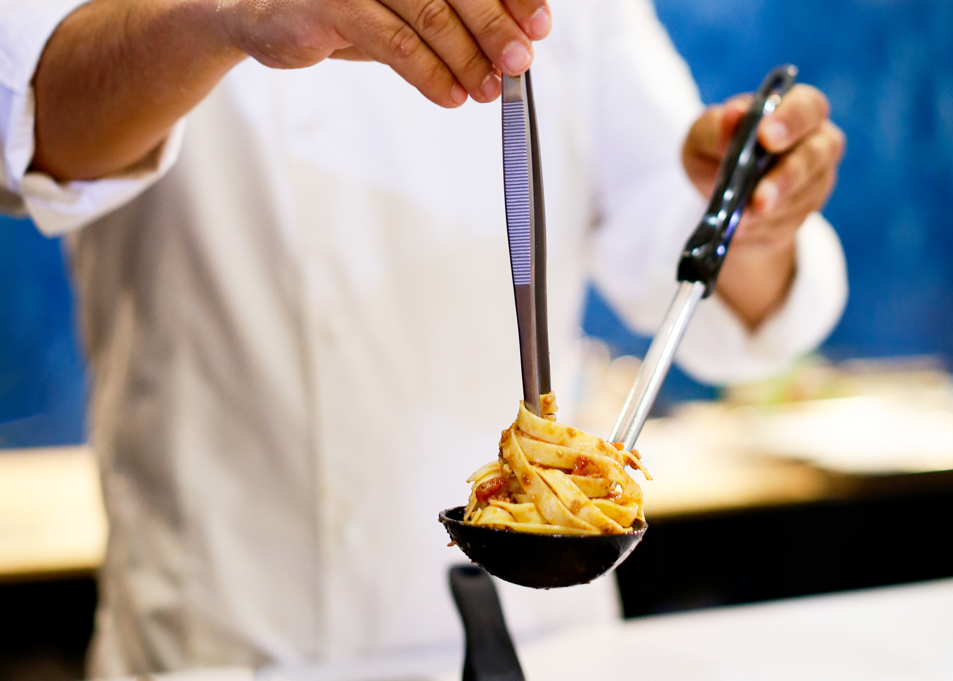 Um chef serve massa com molho de uma concha usando uma pinça.