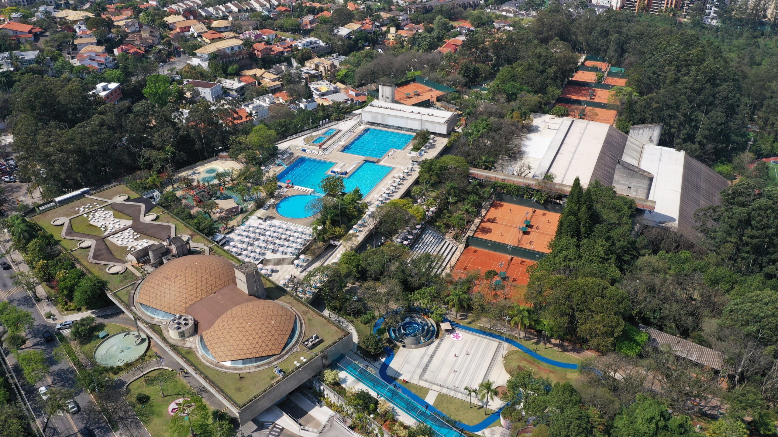 Vista aérea do Clube Paineiras Morumbi, com várias piscinas azuis, quadras de tênis de saibro e extensas áreas verdes. Ao redor, casas e edifícios residenciais se espalham pela paisagem urbana.