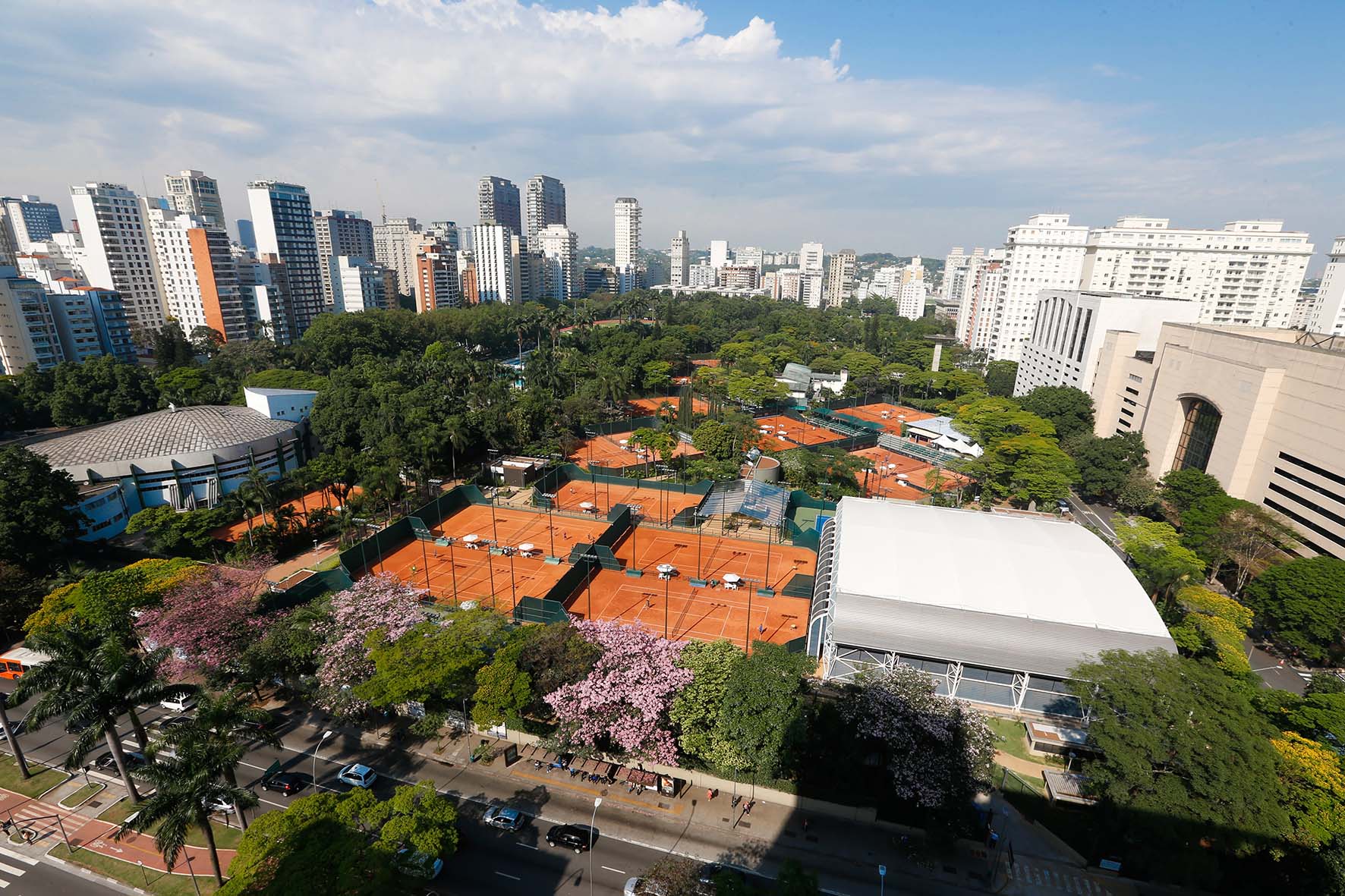 Vista aérea do Clube Pinheiros, em São Paulo, com várias quadras de tênis de saibro e áreas verdes exuberantes, incluindo árvores com flores rosas. Ao redor, edifícios altos de uma cidade se erguem sob um céu azul com nuvens.