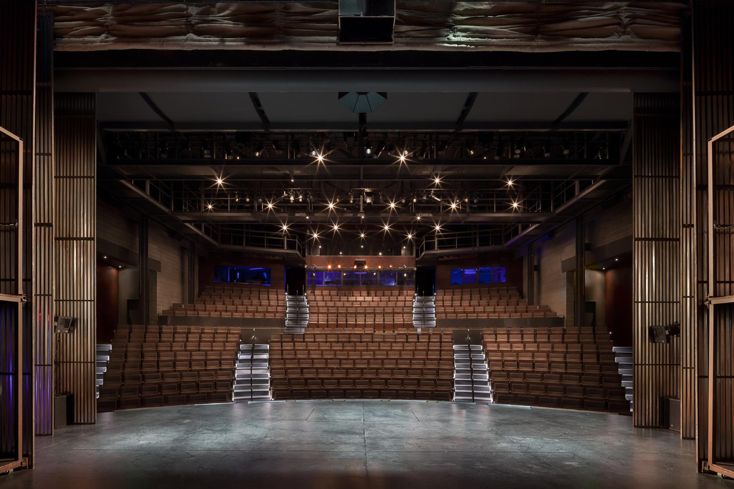 Um teatro moderno com assentos de madeira dispostos em semicírculo, voltados para um palco vazio. Luzes brilhantes no teto iluminam o espaço, e detalhes arquitetônicos em madeira e metal compõem o ambiente.