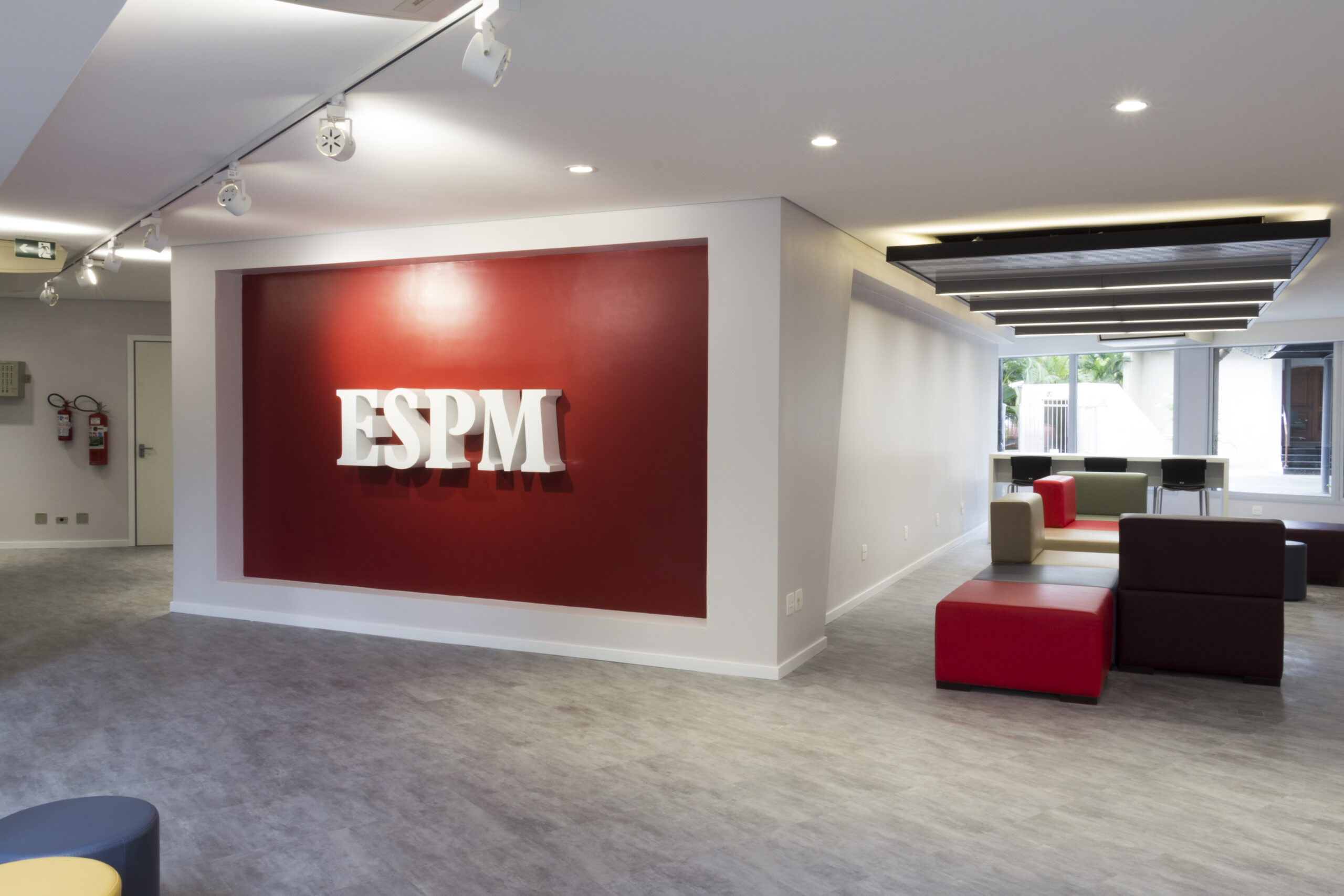 Interior moderno da ESPM campus Vila Mariana com parede vermelha e o logo, além de assentos coloridos e janelas.