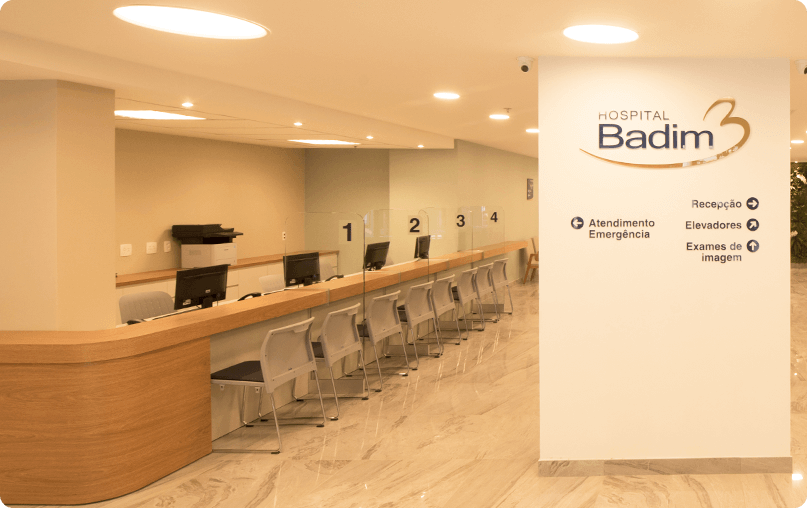 Recepção de hospital moderna e bem iluminada do Hospital Badim, com um balcão de madeira, cadeiras para espera e um painel de sinalização com o logotipo "Hospital Badim".