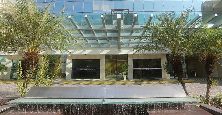 Fachada Hospital Samaritano em Botafogo, com grandes janelas de vidro, entrada coberta e palmeiras. Uma fonte de água com cascata e uma placa metálica complementam a paisagem.