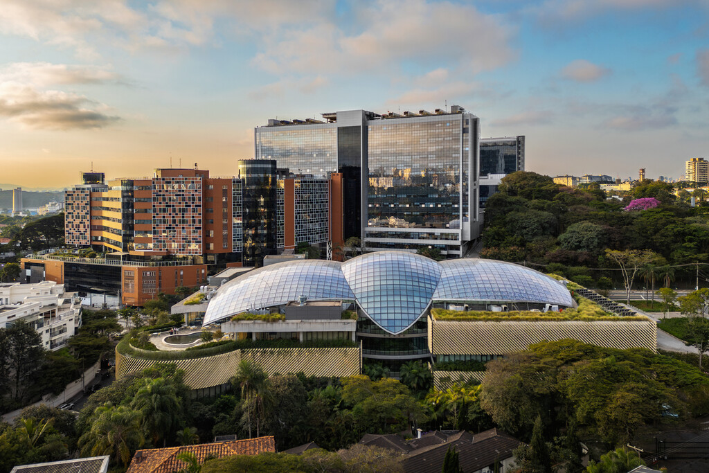 Vista panorâmica do complexo hospitalar Albert Einstein no Morumbi, com telhados de vidro curvos e edifícios altos de fachada espelhada. A paisagem inclui áreas verdes e outros prédios urbanos sob um céu parcialmente nublado.