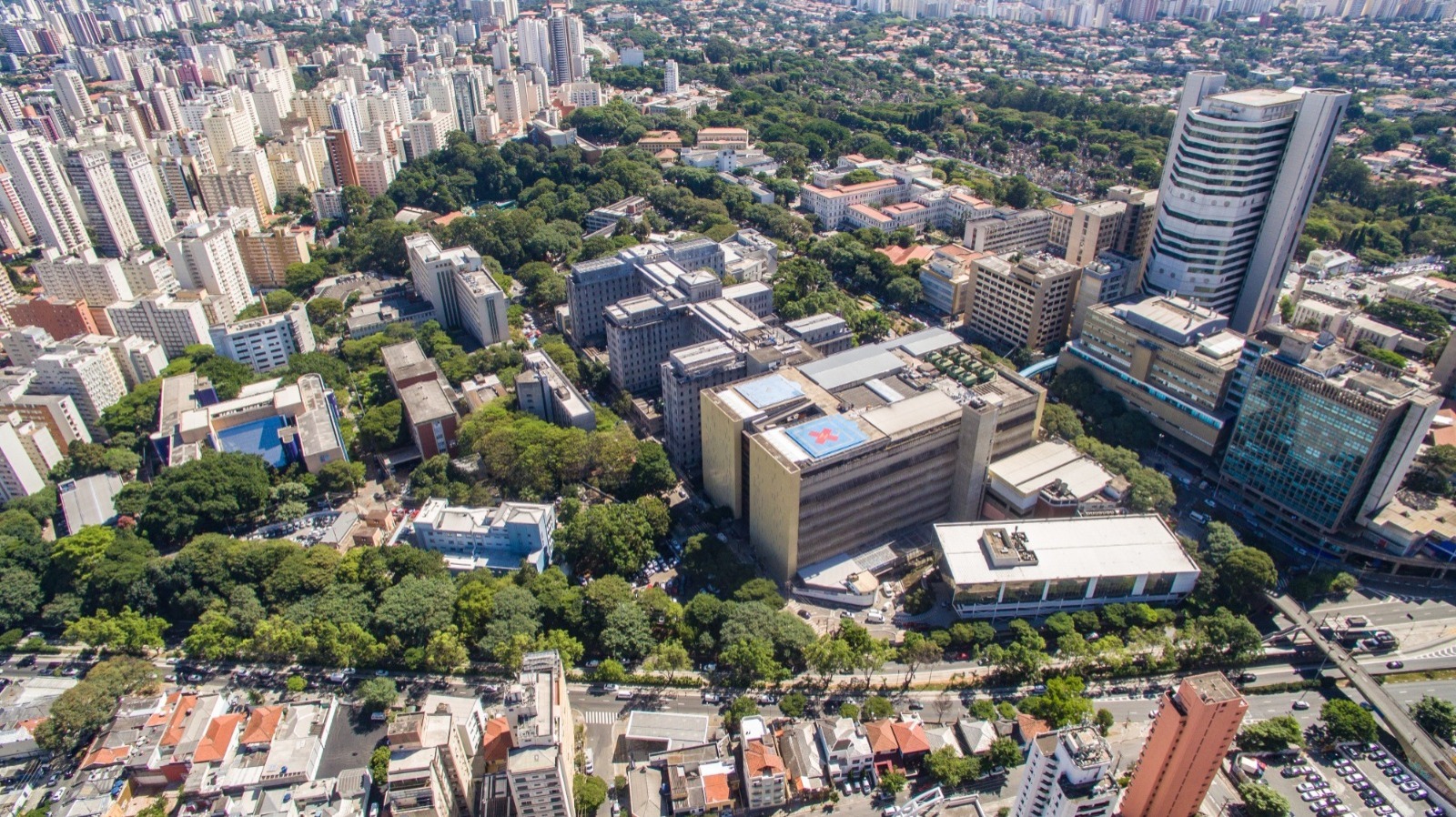 Vista aérea do complexo hospitalar do Hospital das Clínicas, em São Paulo, com múltiplos edifícios de diferentes alturas e estilos, cercado por áreas verdes. A paisagem urbana densa de São Paulo se estende ao fundo, com muitos prédios residenciais.