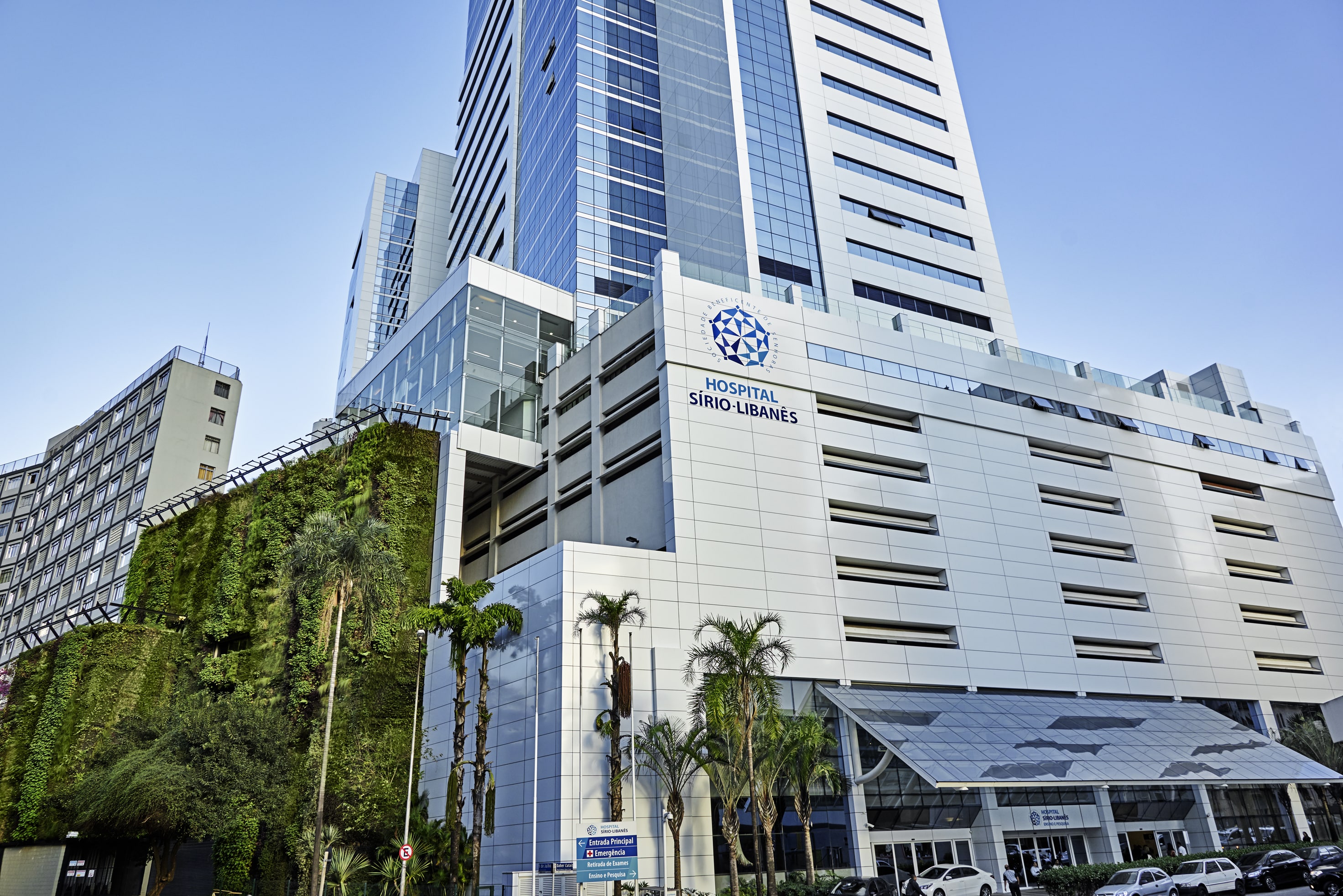 Fachada moderna do Hospital Sírio-Libanês, com um grande painel verde vertical e palmeiras na entrada. O edifício de vidro e concreto reflete o céu azul, e carros estão estacionados em frente.