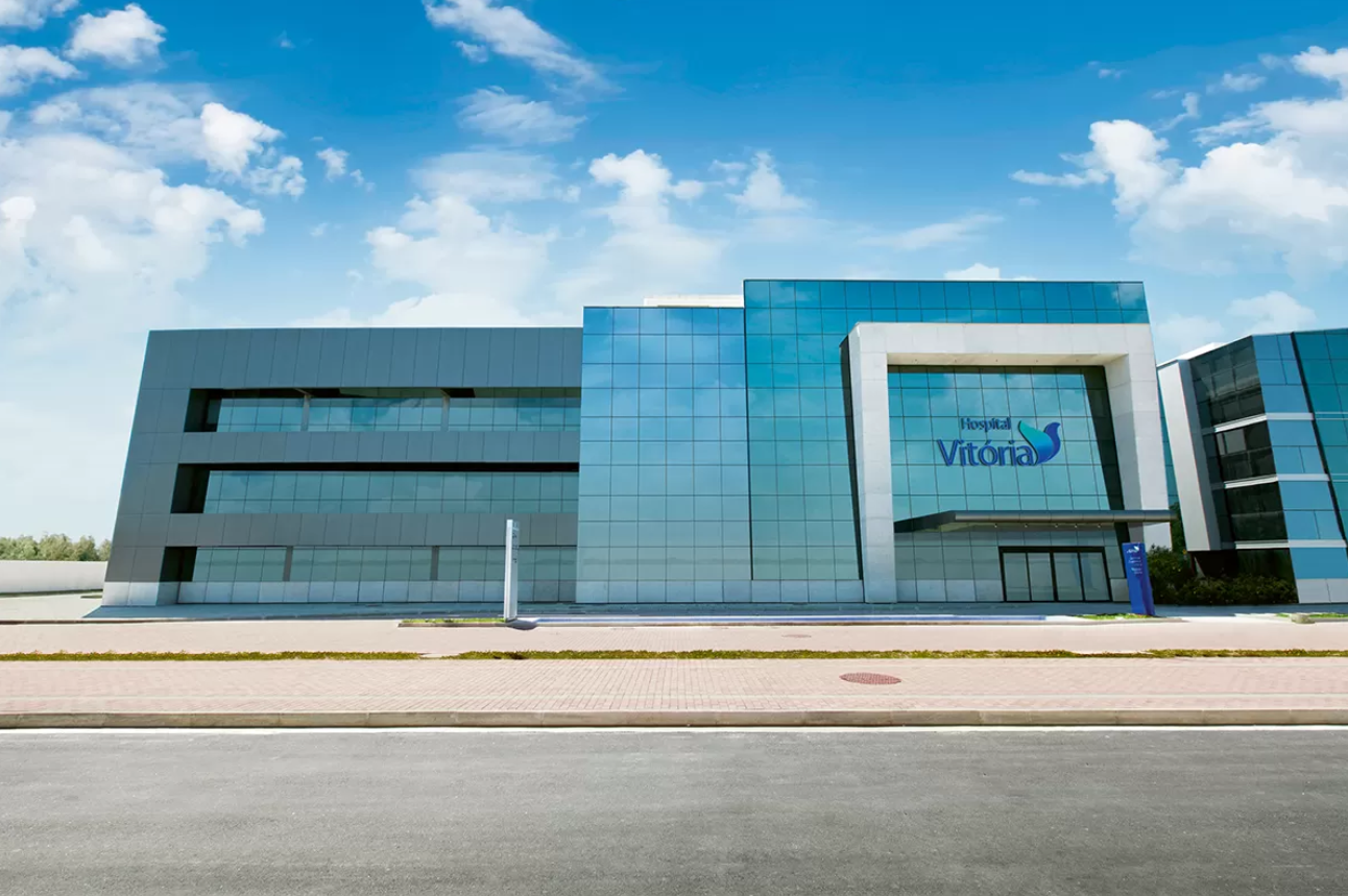 Um edifício moderno com fachada de vidro azul e cinza, com o logotipo "Hospital Vitória" em destaque, sob um céu azul com nuvens.