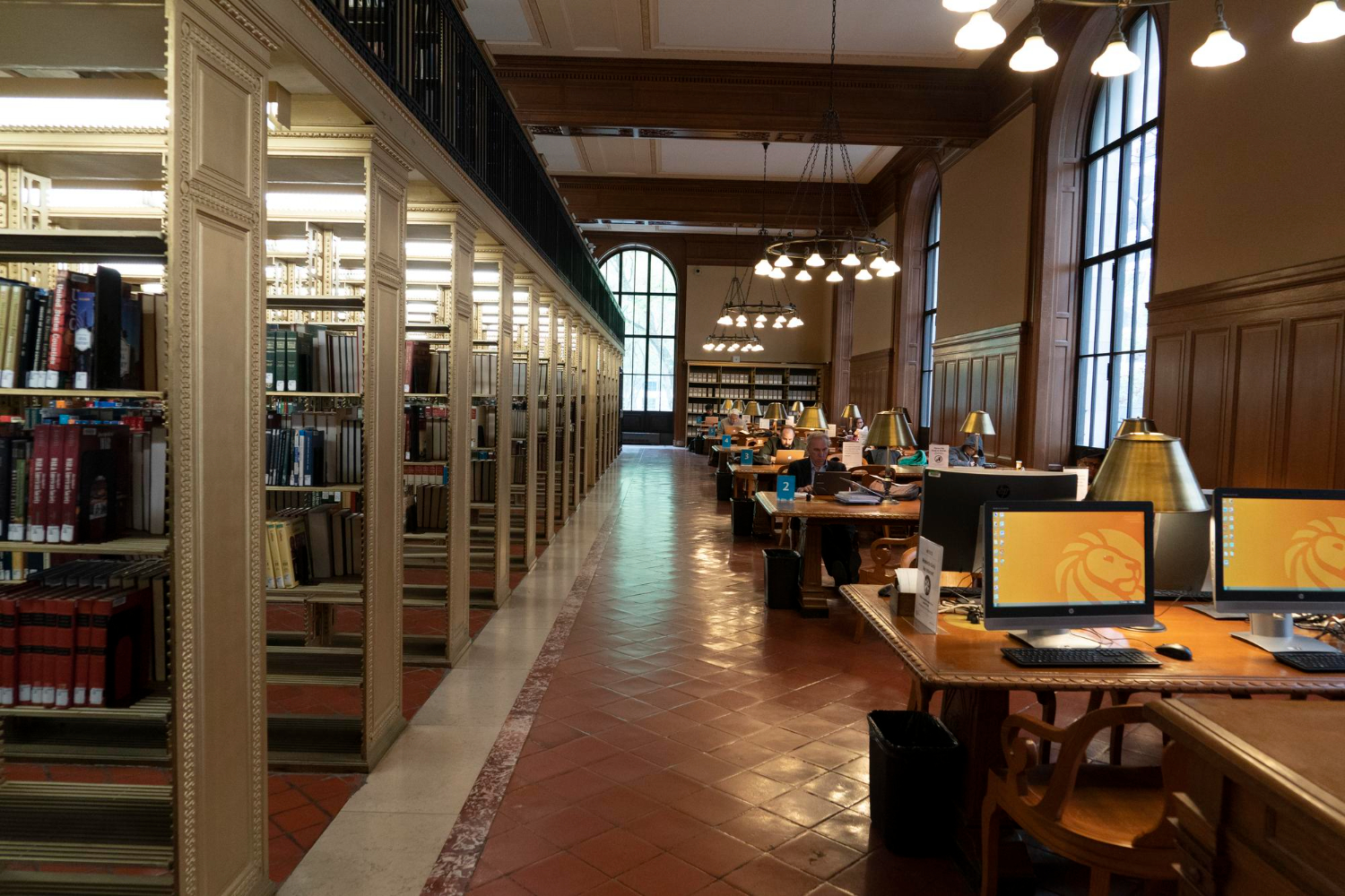 Um corredor de biblioteca com estantes de madeira repletas de livros à esquerda e mesas de estudo com computadores à direita. A iluminação aconchegante e as grandes janelas criam um ambiente propício à leitura.