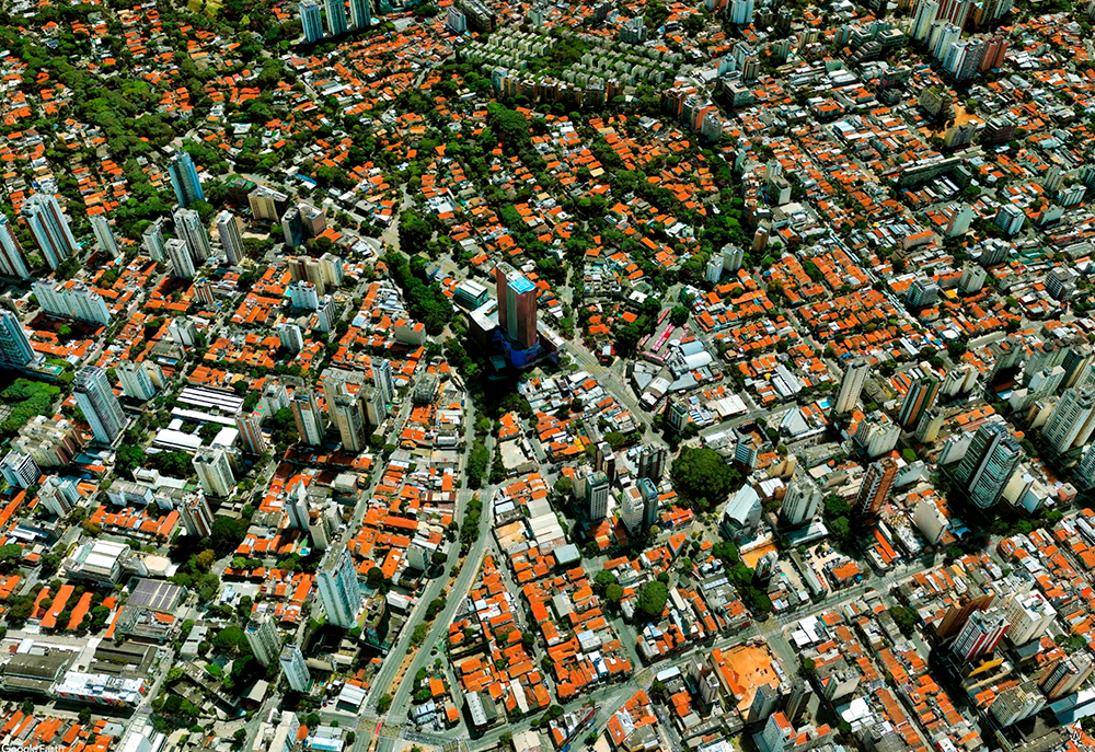 Vista aérea de uma densa área urbana com muitos edifícios e telhados alaranjados, intercalados por manchas verdes de árvores. Ruas e avenidas serpenteiam pela paisagem, e o edifício do Instituto Tomie Ohtake se destaca no centro.