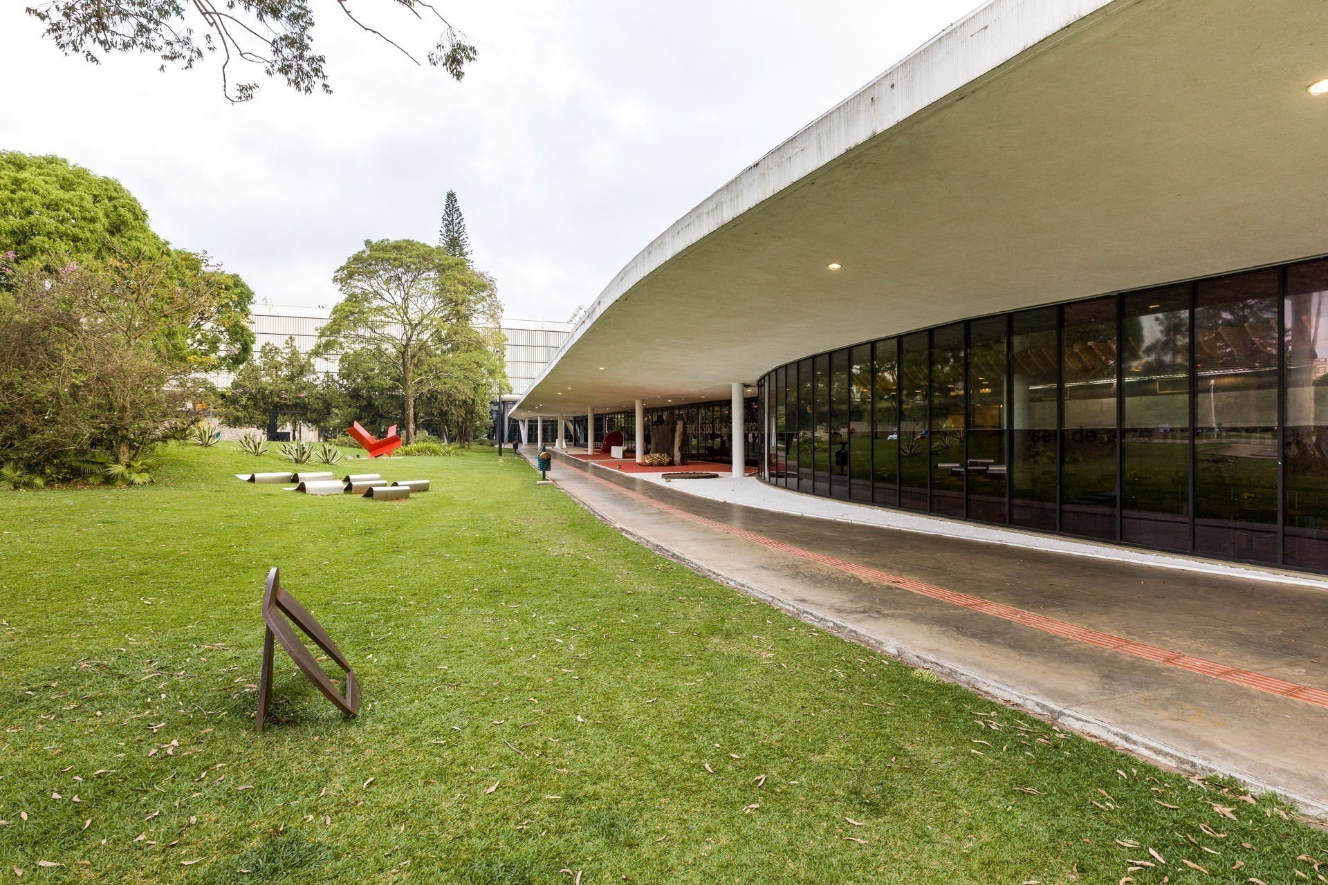 Vista externa do Museu de Arte Moderna (MAM) em São Paulo, com sua arquitetura curva e envidraçada, cercada por um gramado verde com esculturas e árvores.