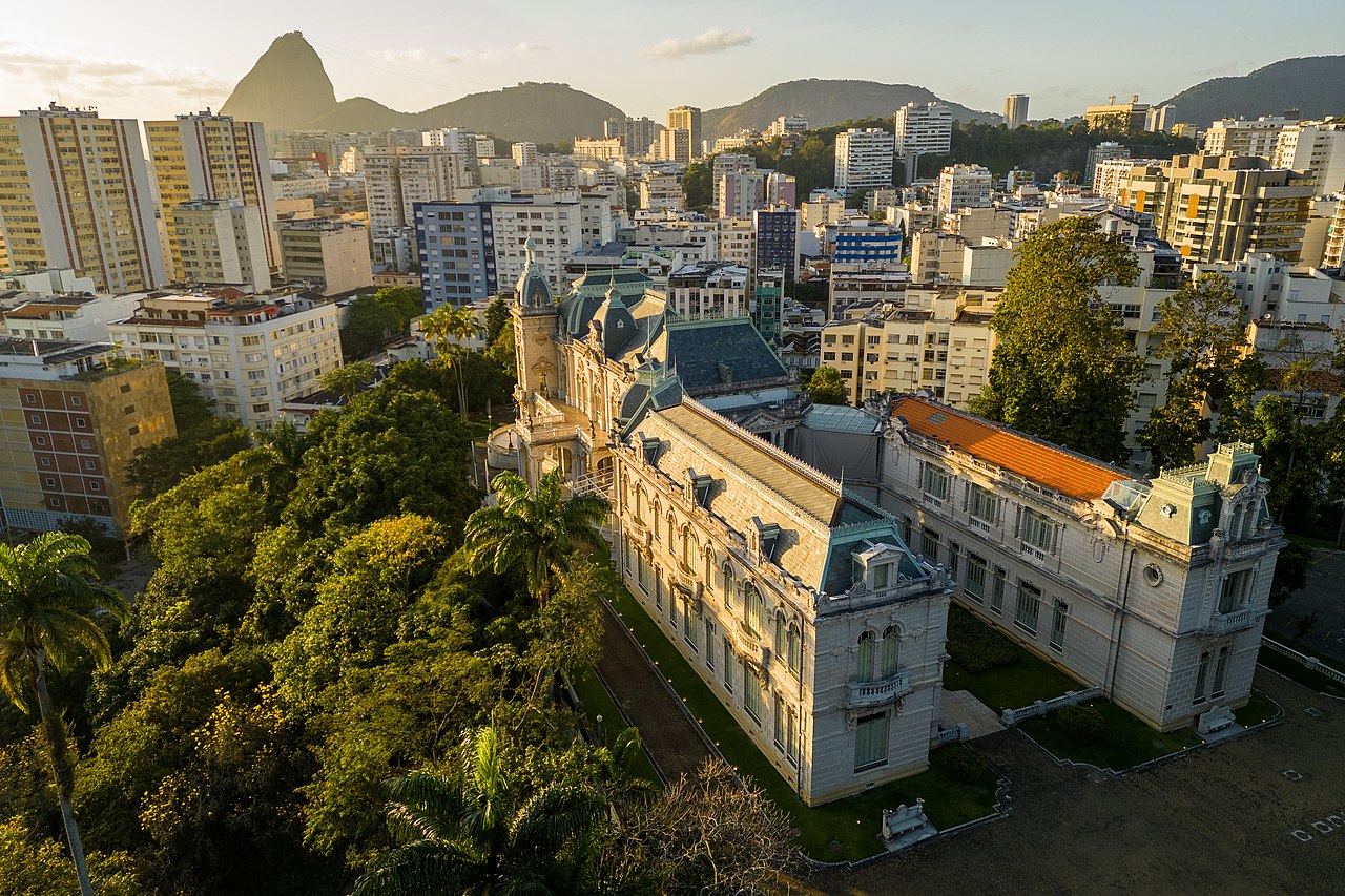 Vista aérea do bairro Laranjeira, focando no Palácio Guanabara, com telhados verdes e laranjas, cercado por árvores exuberantes e a paisagem urbana do Rio de Janeiro ao fundo, com montanhas e edifícios.