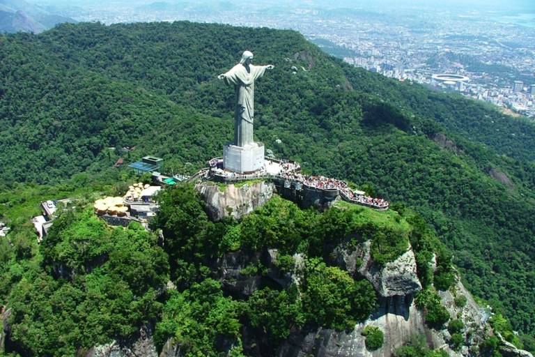 Vista aérea do Parque Nacional Tijuca, com foco no Cristo Redentor no topo de uma montanha, cercado por densa vegetação e com a cidade do Rio de Janeiro ao fundo.