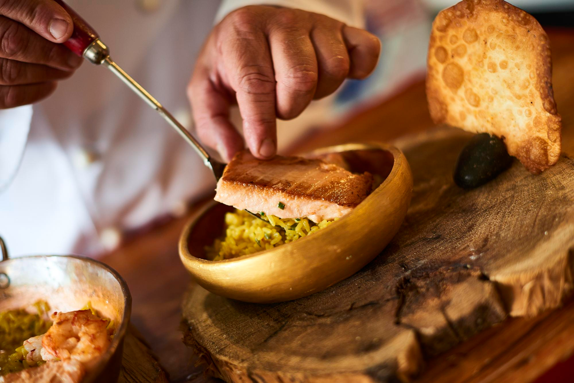 Um chef finalizando um prato gourmet, colocando um pedaço de salmão grelhado sobre arroz amarelo em uma tigela de madeira, com um pastel e um abacate ao lado.