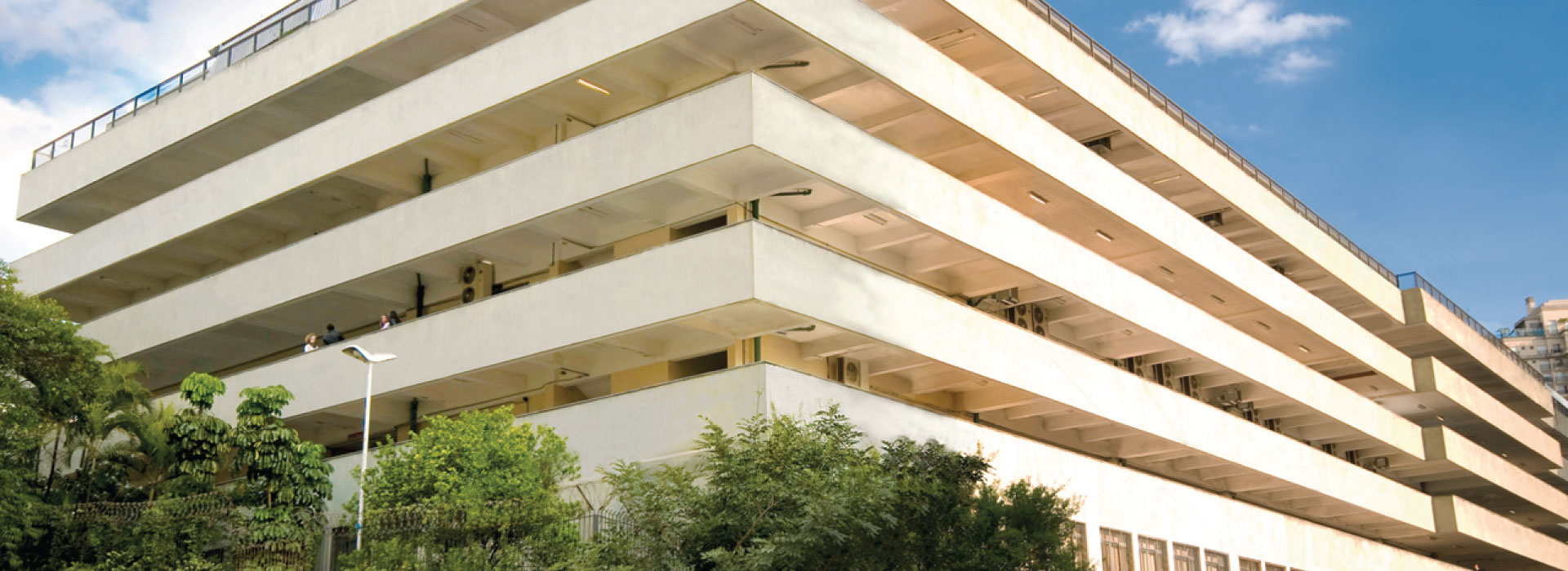 Edifício PUC Perdizes com múltiplos andares com uma arquitetura em camadas e balcões salientes, cercado por árvores verdes. O céu azul com algumas nuvens é visível ao fundo, e a estrutura se destaca pela sua imponência.