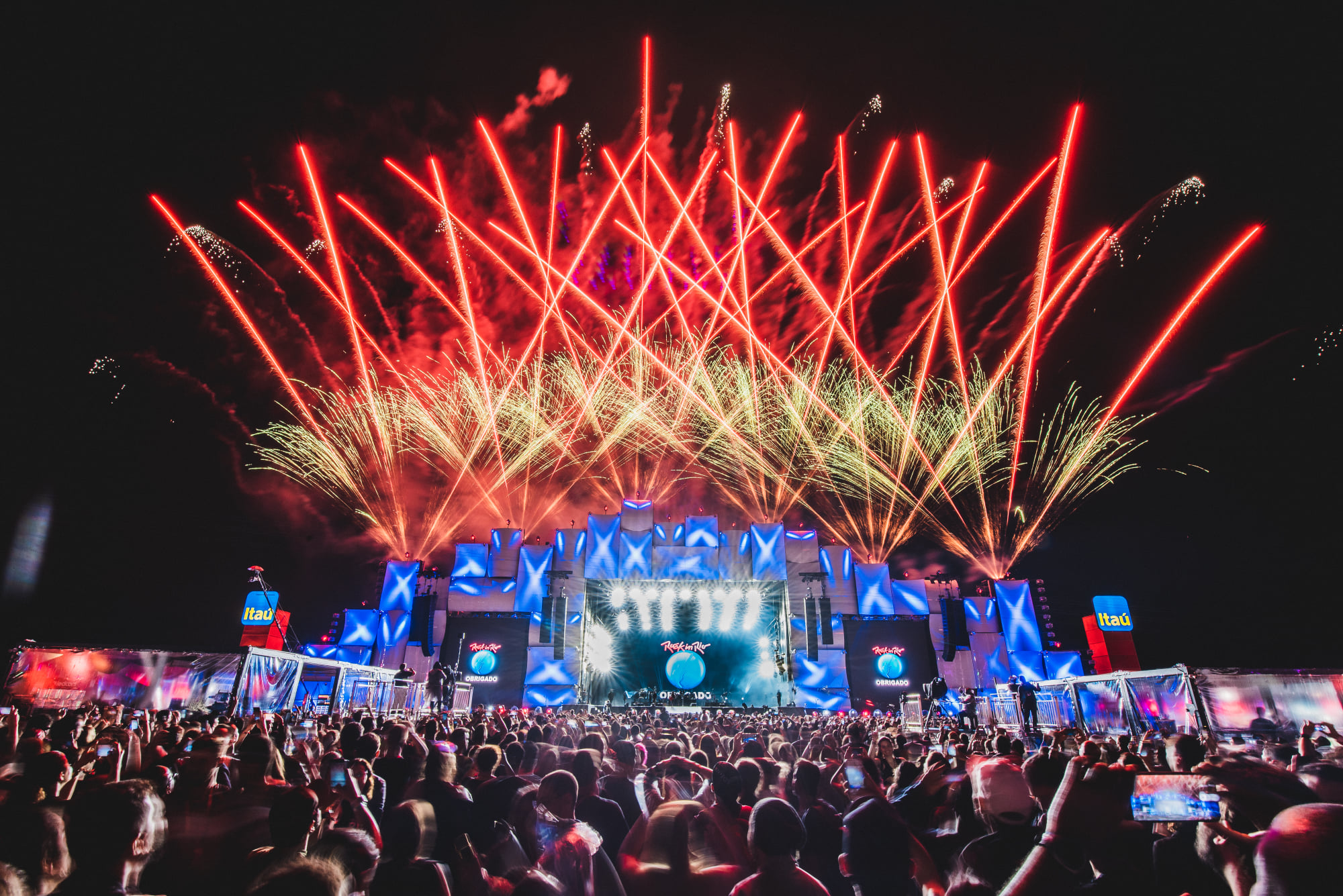 Festival Rock in Rio. Fogos de artifício vermelhos e dourados iluminam o céu noturno acima de um palco de festival de música com luzes azuis e uma multidão de pessoas.