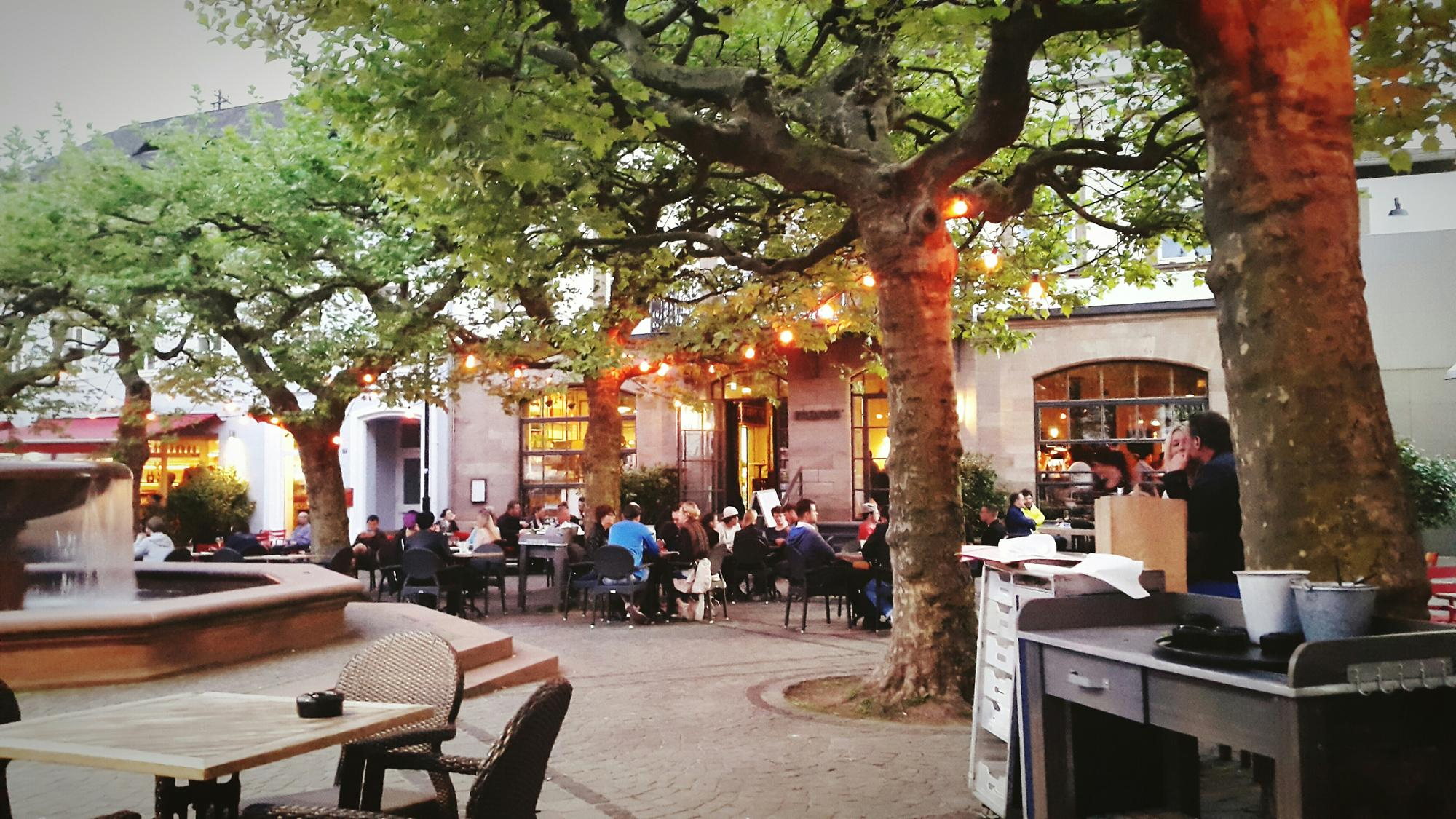 Uma praça charmosa com mesas de restaurante ao ar livre sob a sombra de grandes árvores com luzes penduradas. Uma fonte de pedra e pessoas desfrutando da noite criam uma atmosfera acolhedora.