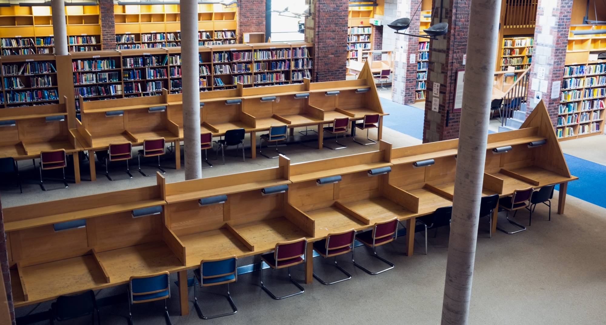 Sala de estudos de biblioteca com mesas de madeira individuais e cadeiras coloridas. Estantes repletas de livros ao fundo, com pilares e paredes de tijolos.