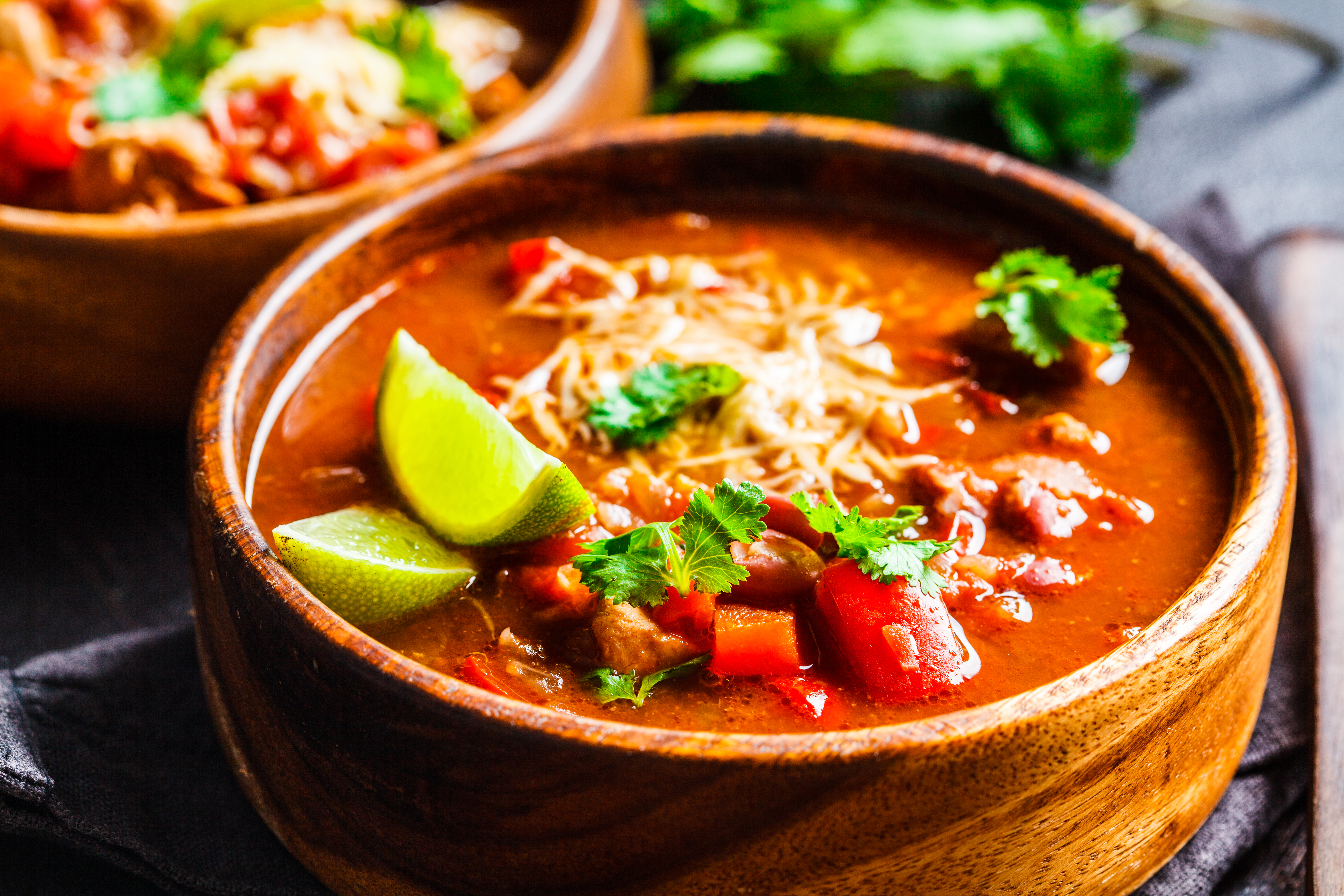 Close-up de uma tigela de madeira com sopa de feijão mexicana. A sopa avermelhada contém tomate, feijão, queijo ralado, coentro fresco e fatias de limão-siciliano.