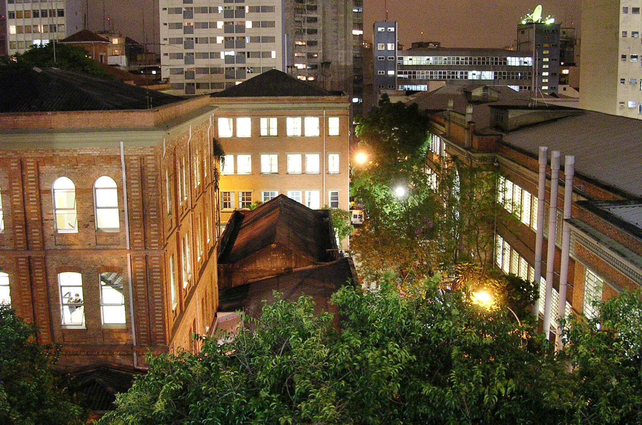 Vista noturna de edifícios antigos da Universidade Mackenzie, com janelas iluminadas e telhados escuros. Árvores e postes de luz adicionam profundidade à cena, enquanto prédios mais modernos se elevam ao fundo.
