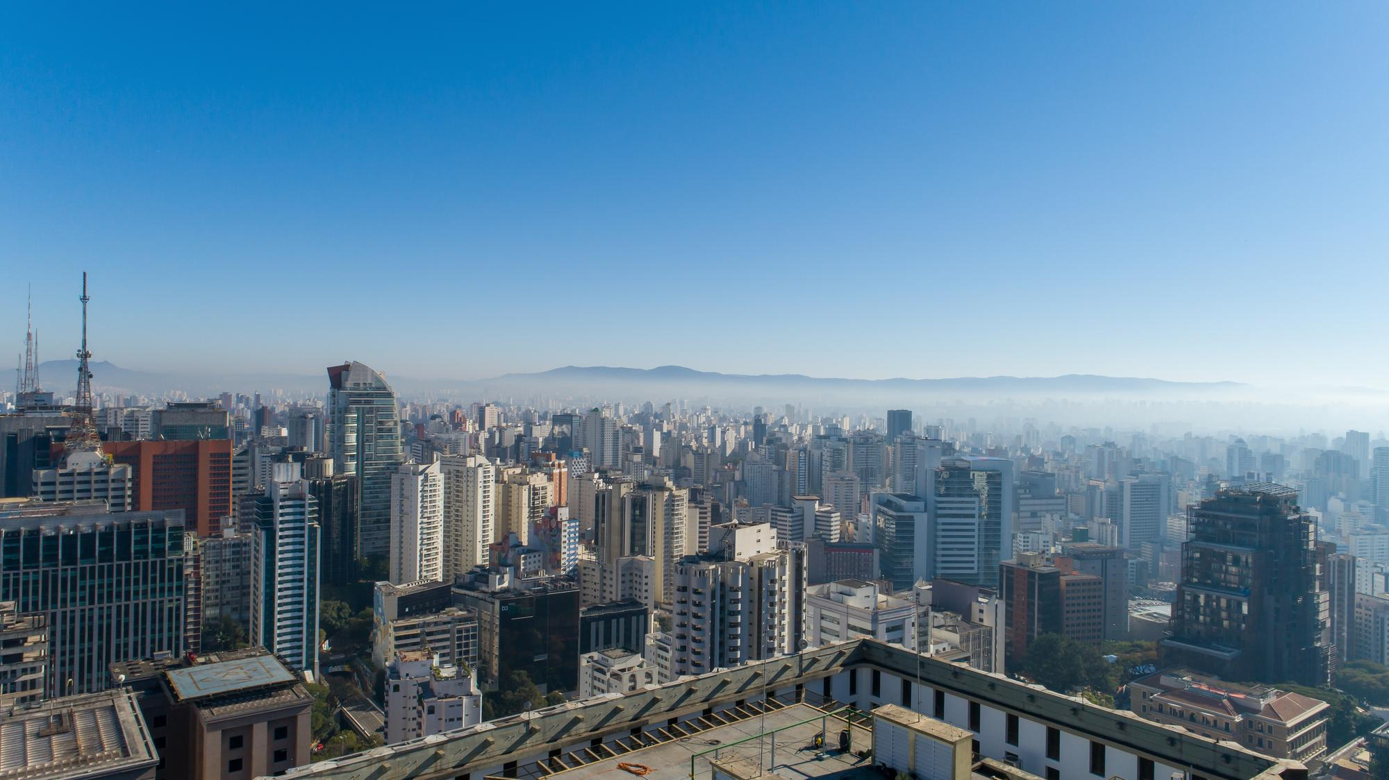 Vista aérea de uma vasta paisagem urbana com inúmeros edifícios de diferentes alturas e estilos, estendendo-se até o horizonte. Uma leve névoa cobre a cidade ao longe, e montanhas são visíveis sob um céu azul claro.