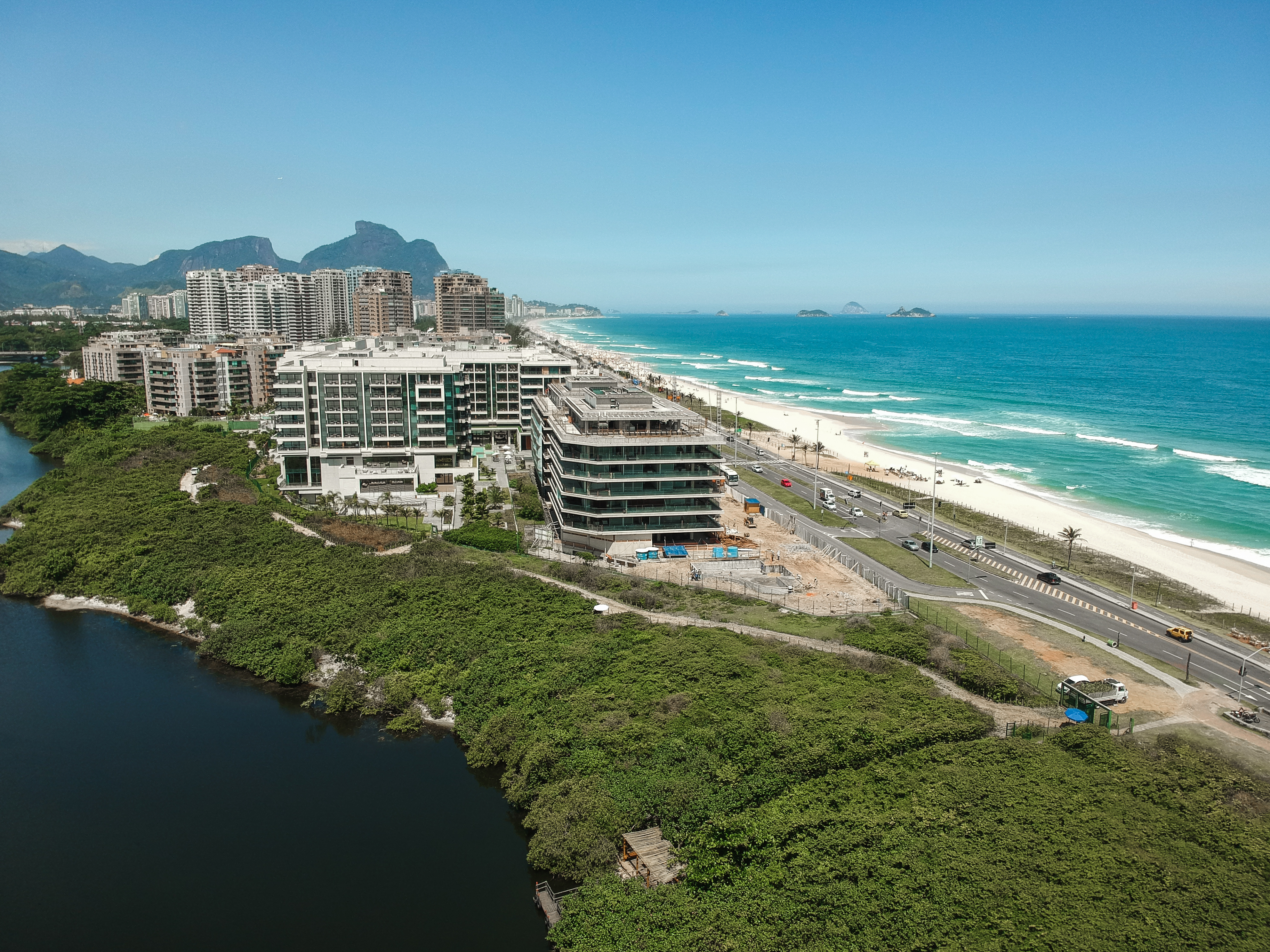 Vista aérea da Praia da Reserva, com um lago e vegetação densa em primeiro plano, prédios e uma estrada movimentada na orla, e o oceano azul com ondas quebrando na praia.