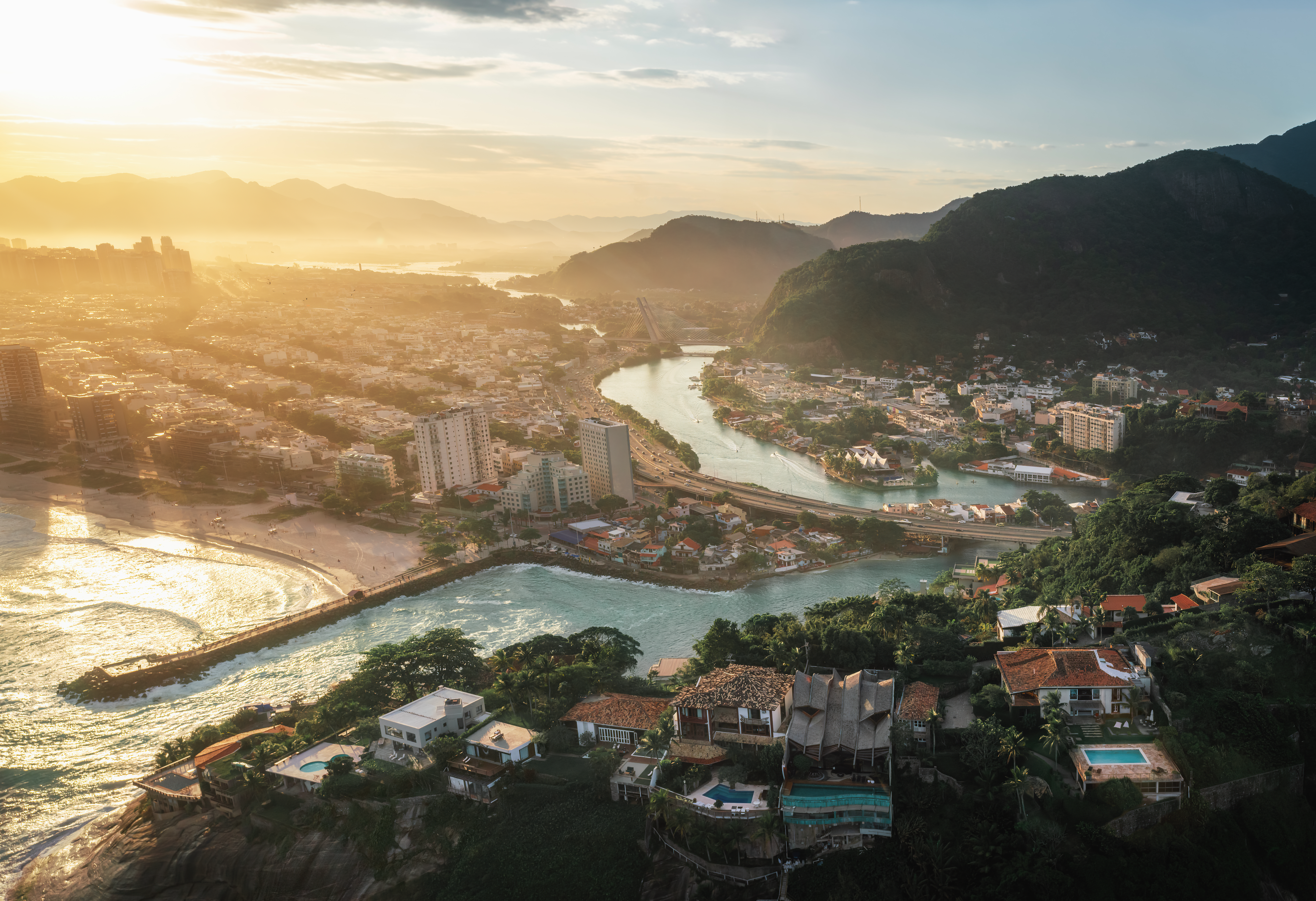 Vista aérea da Barra da Tijuca, com um rio serpenteando por uma área urbana, uma praia com ondas quebrando, e montanhas ao fundo sob um céu dourado de pôr do sol.