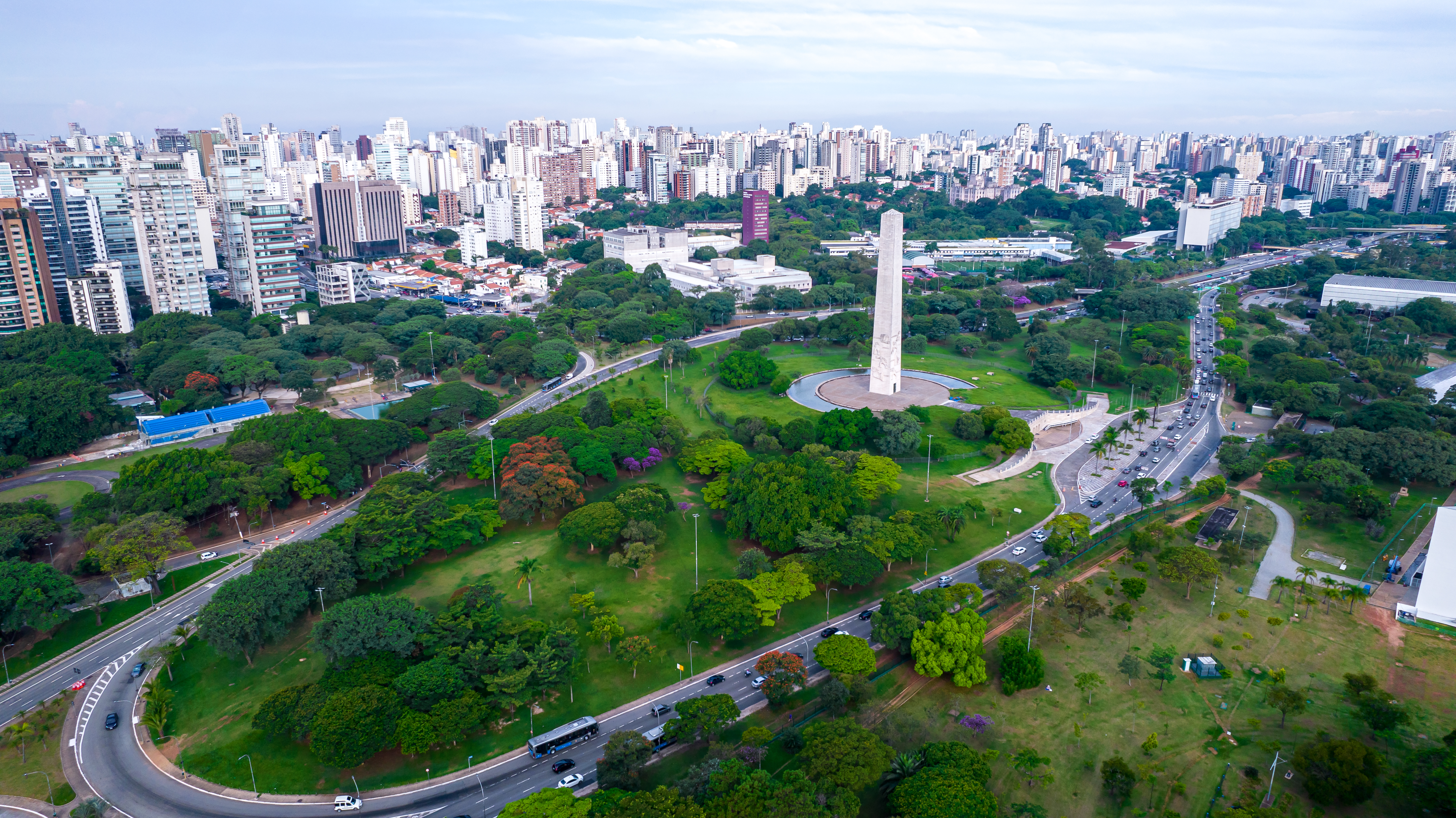 Vista aérea do Parque Ibirapuera em São Paulo, com o Obelisco do Ibirapuera em destaque, cercado por áreas verdes e edifícios residenciais ao fundo.