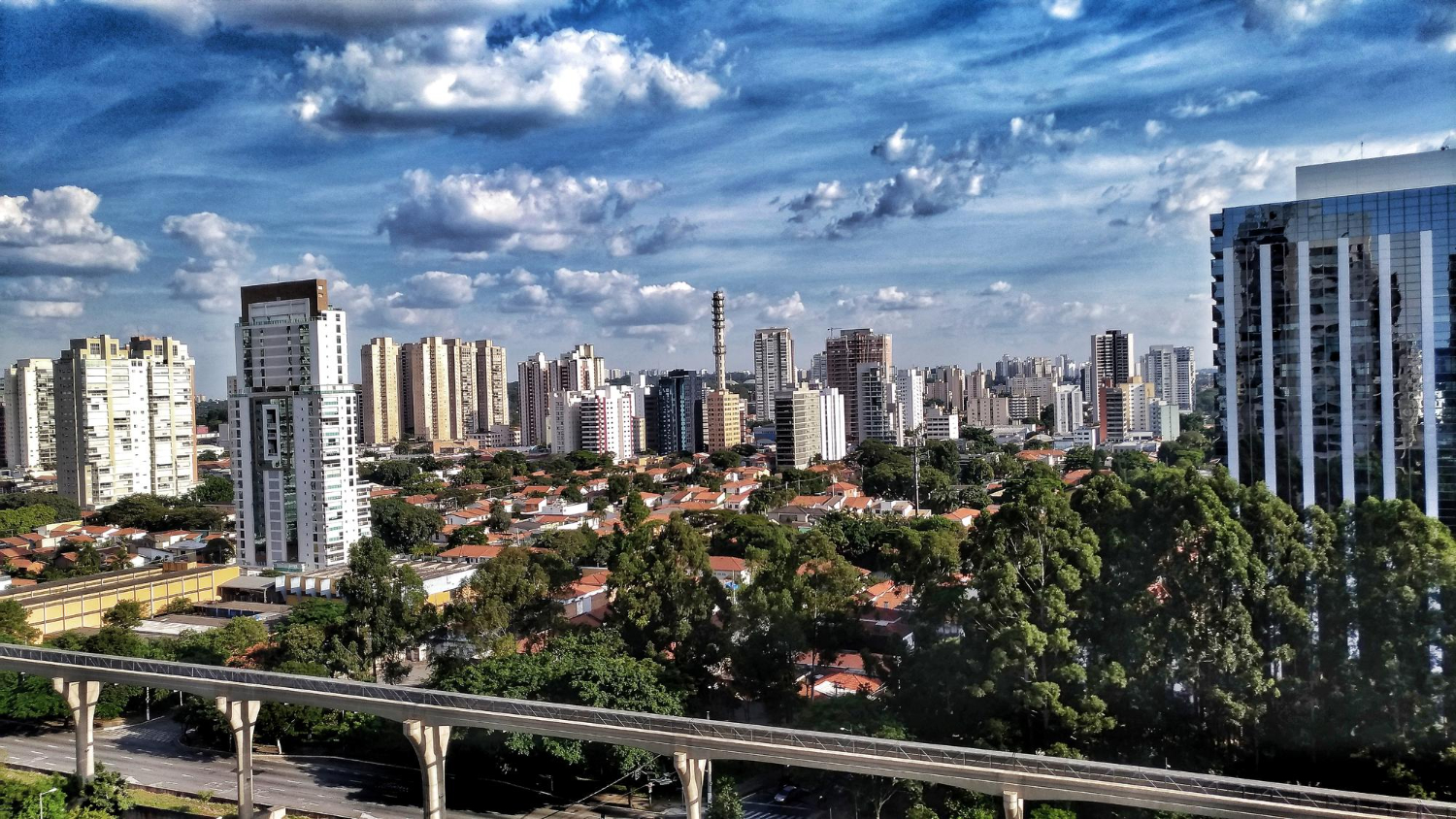 Vista panorâmica de uma cidade com uma mistura de edifícios altos e casas com telhados de terracota, cercadas por árvores. Um trilho elevado atravessa a paisagem, e o céu azul está repleto de nuvens brancas.