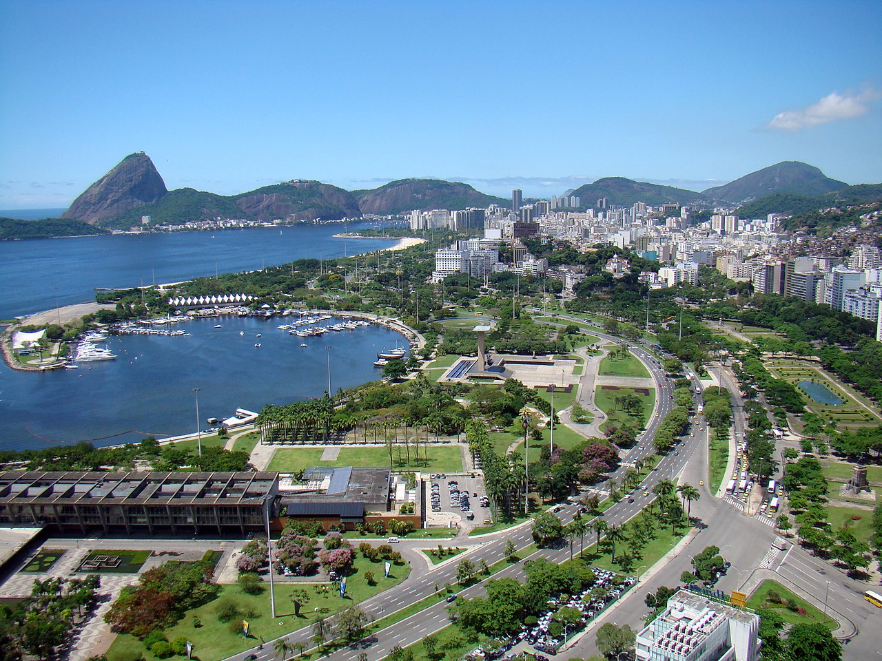 Vista aérea da Marina da Glória no Rio de Janeiro, com barcos na baía, o Pão de Açúcar ao fundo, e a cidade se estendendo com áreas verdes e estradas.