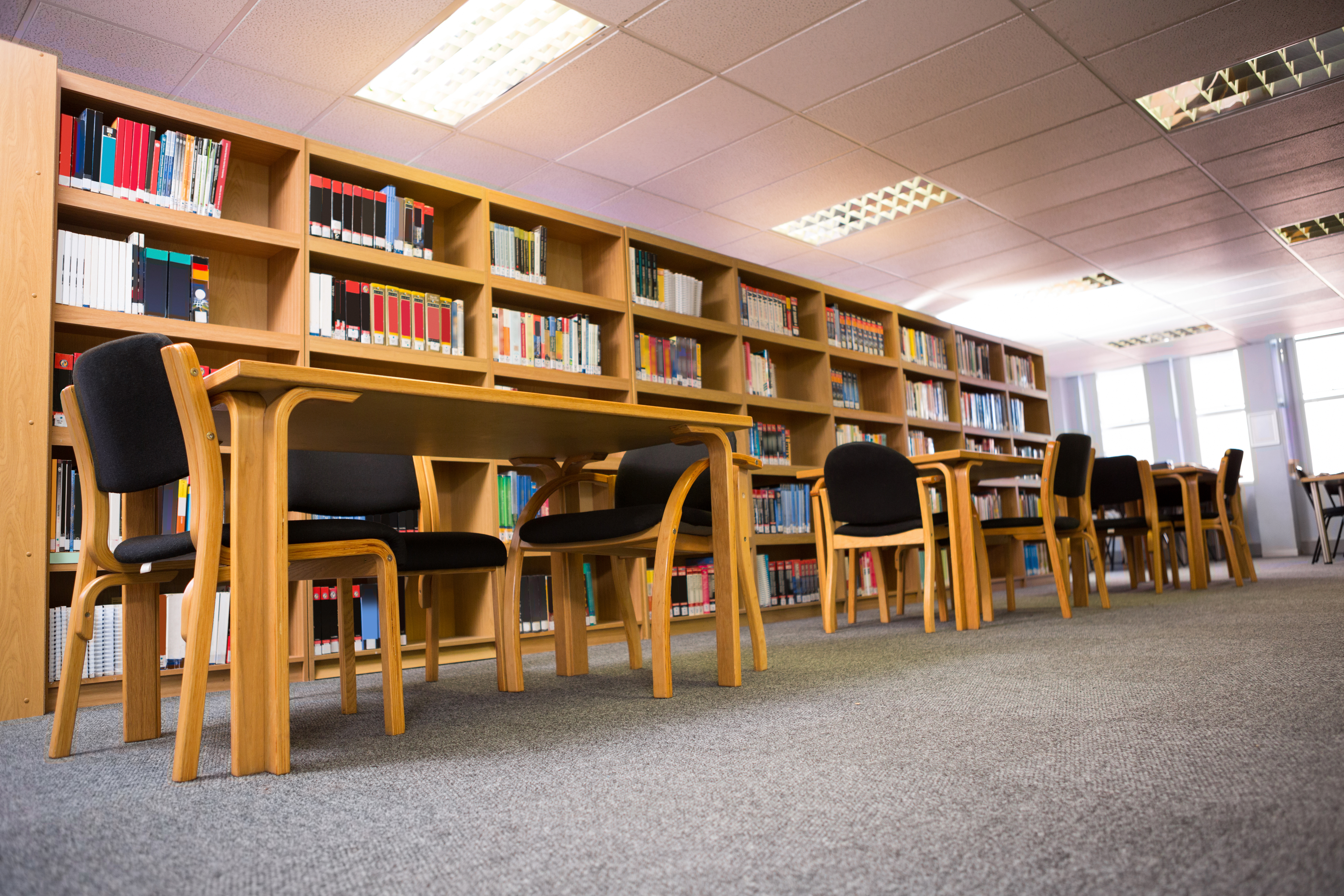 Uma biblioteca com estantes de madeira cheias de livros coloridos, mesas de madeira e cadeiras pretas dispostas em fileiras, e um carpete cinza no chão.