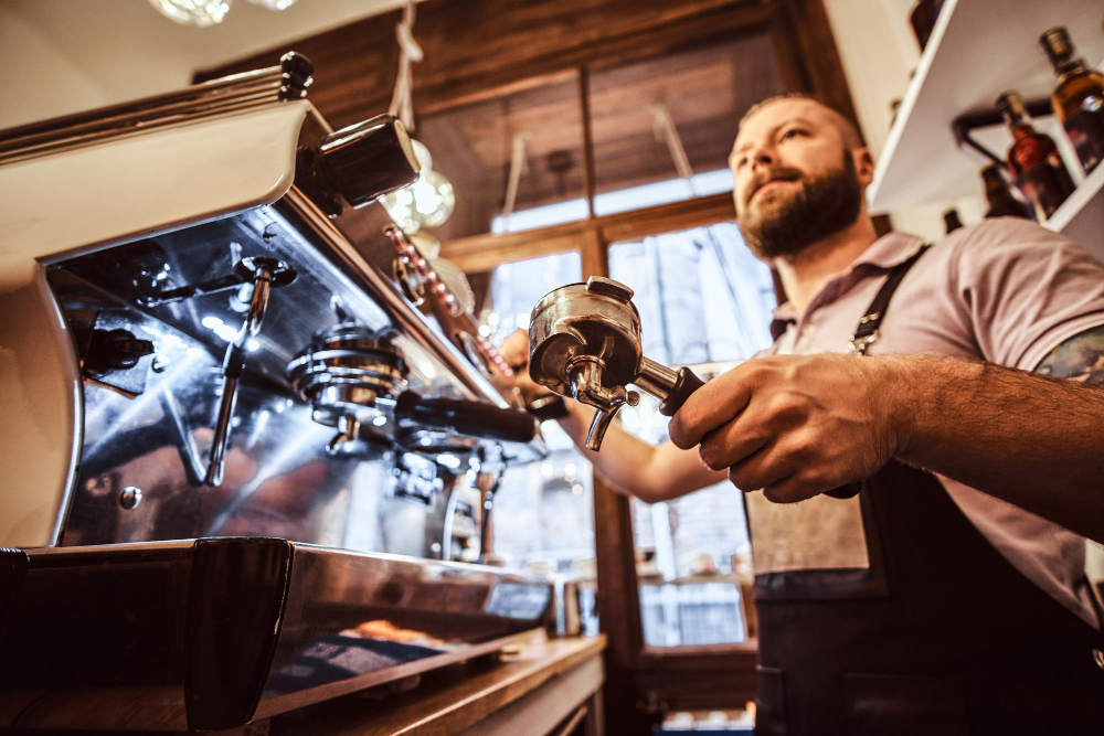 Um barista barbudo com um avental preto opera uma máquina de café expresso de metal, segurando um porta-filtro e olhando para cima, em uma cafeteria.