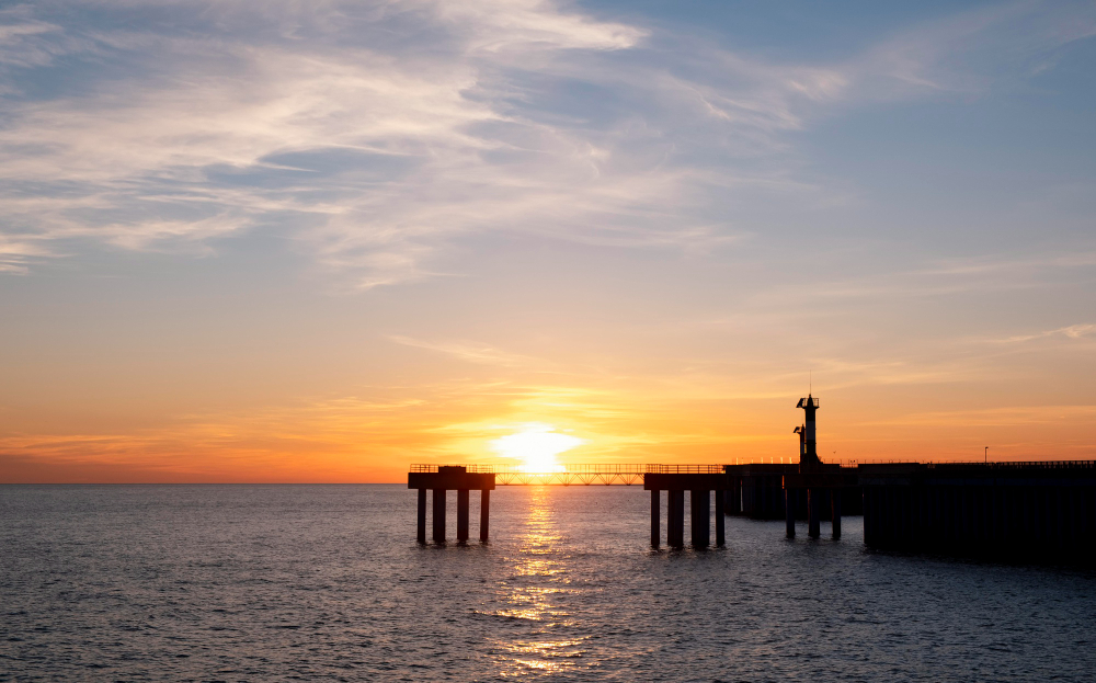 Um pôr do sol vibrante sobre o oceano, com o sol laranja refletindo na água, e a silhueta de um cais com pilares e uma torre ou farol ao longe.