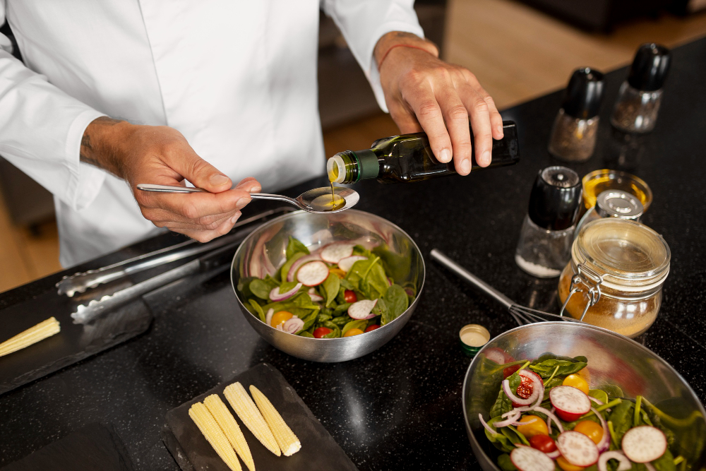 Um chef em um jaleco branco derramando azeite de uma garrafa em uma colher sobre uma tigela de salada fresca com espinafre, rabanetes e tomate cereja em uma bancada preta.