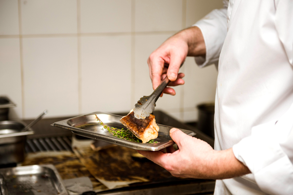 Um chef com um jaleco branco usa uma pinça para transferir um pedaço de peixe grelhado com ervas de uma grelha para uma bandeja de metal.