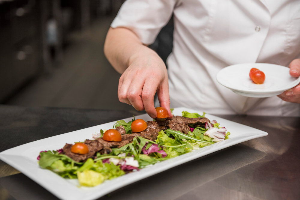Um chef com tatuagens nos braços, vestindo um jaleco branco, adiciona molho de uma tigela de vidro a uma salada fresca em uma tigela de metal, em uma bancada preta com outros ingredientes.