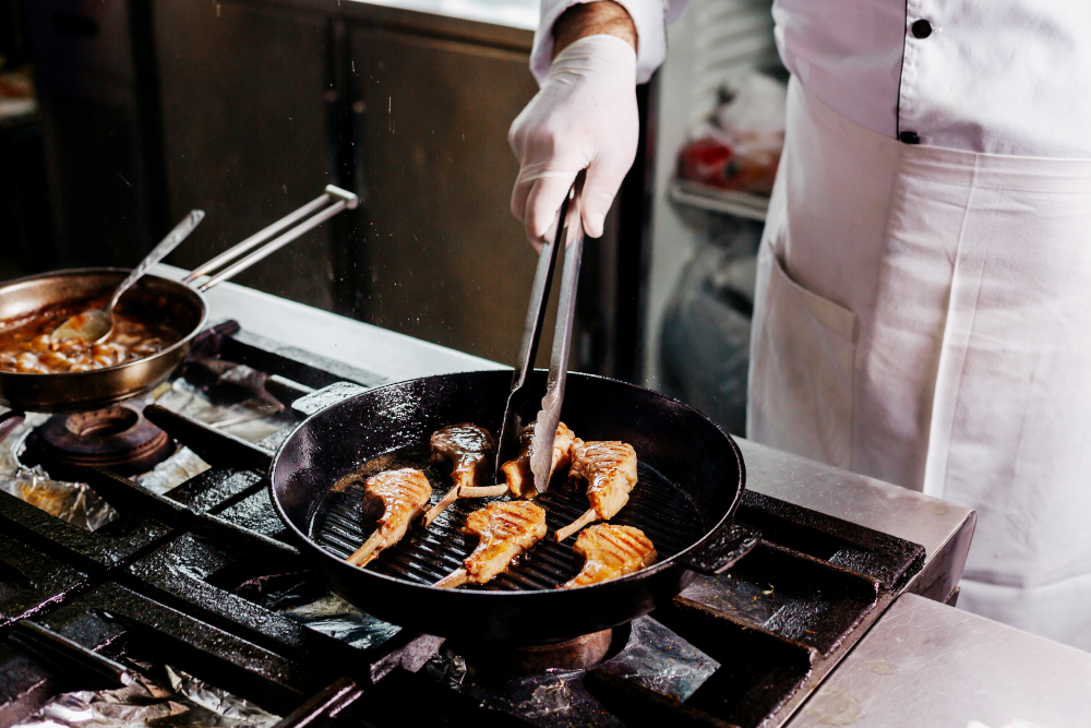 Um chef com luvas brancas usa uma pinça para virar costeletas de cordeiro grelhadas em uma frigideira preta sobre um fogão industrial.