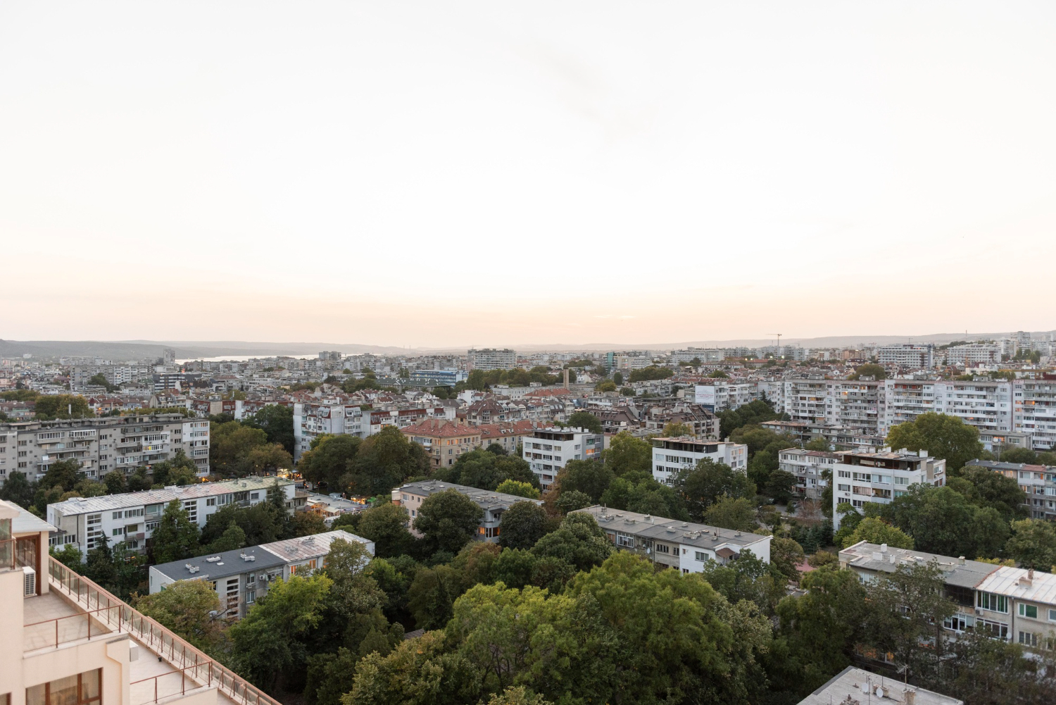 Vista aérea panorâmica de uma cidade densa com edifícios residenciais, muitas árvores verdes e um corpo de água ao fundo, sob um céu claro e pálido.