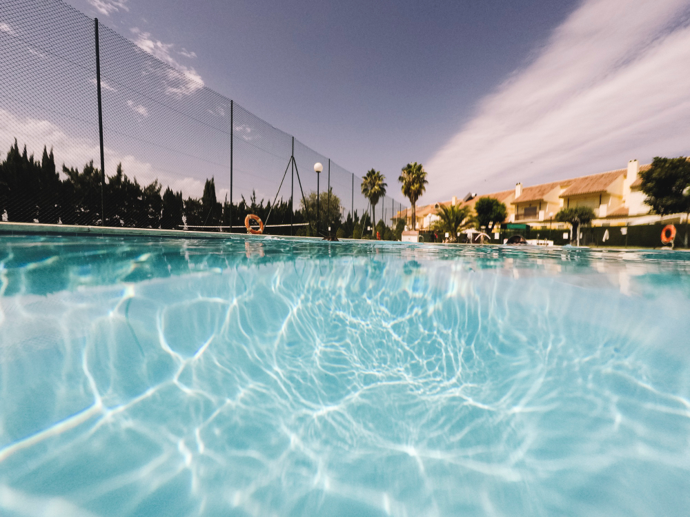 Uma vista de uma piscina com água azul clara e reflexos de luz, com a linha d'água dividindo a imagem. Acima da água, há uma cerca, árvores, palmeiras e edifícios ao fundo sob um céu azul.