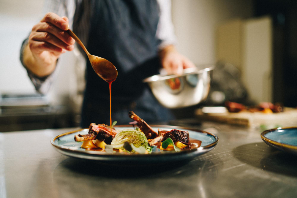 Um chef em um avental está finalizando um prato gourmet, regando molho com uma colher sobre carne, um vegetal verde e purê em um prato azul, em uma cozinha profissional.