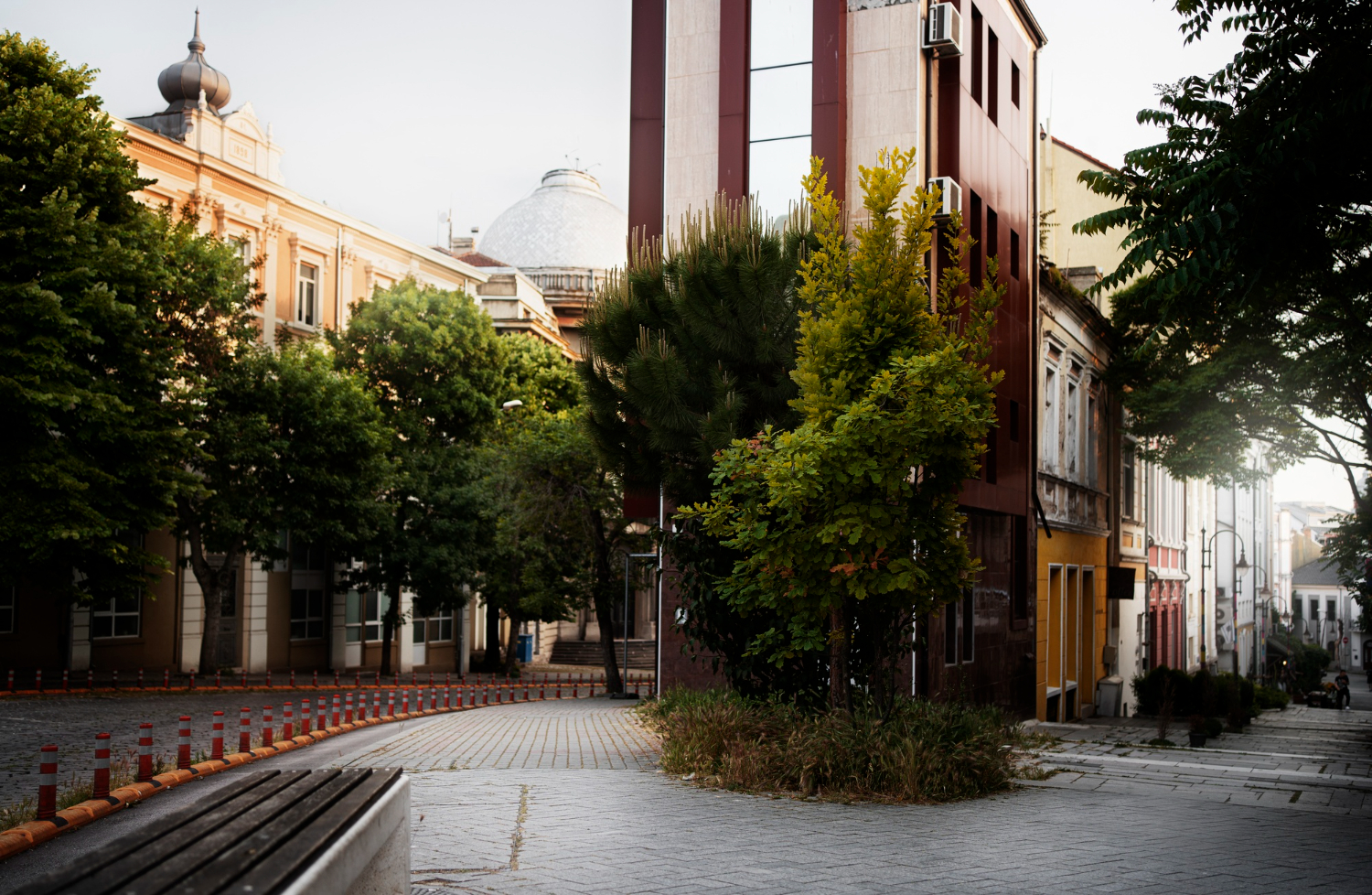Uma rua urbana com edifícios de arquitetura variada, árvores verdes exuberantes, um banco de parque em primeiro plano e uma rua curva com bollards, sob um céu claro.