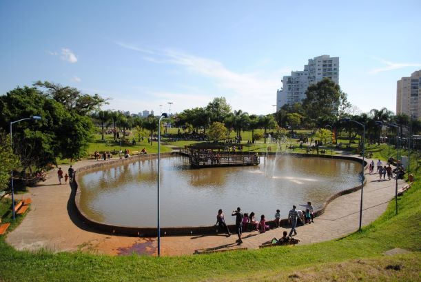 Vista panorâmica de um parque urbano com um lago central, um deck de madeira e uma fonte, cercado por grama verde, árvores e pessoas caminhando e relaxando, com edifícios altos ao fundo sob um céu azul.