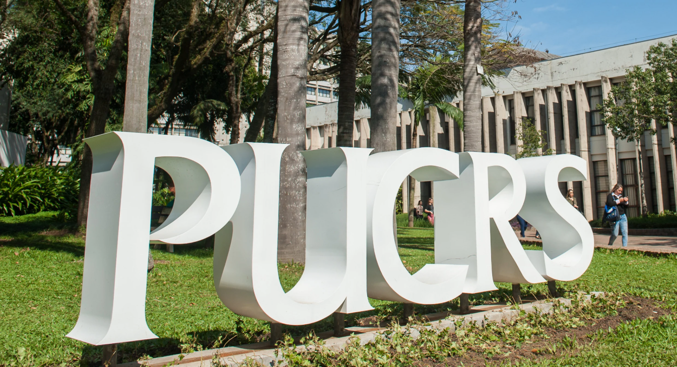 Um grande letreiro branco com as letras "PUCRS" em um gramado verde, com árvores e um edifício universitário moderno ao fundo.