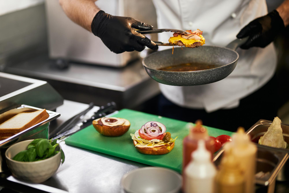 Um chef com luvas pretas usa uma pinça para transferir um hambúrguer com queijo e bacon de uma frigideira para um pão de hambúrguer com alface, tomate e cebola roxa em uma tábua de corte verde.