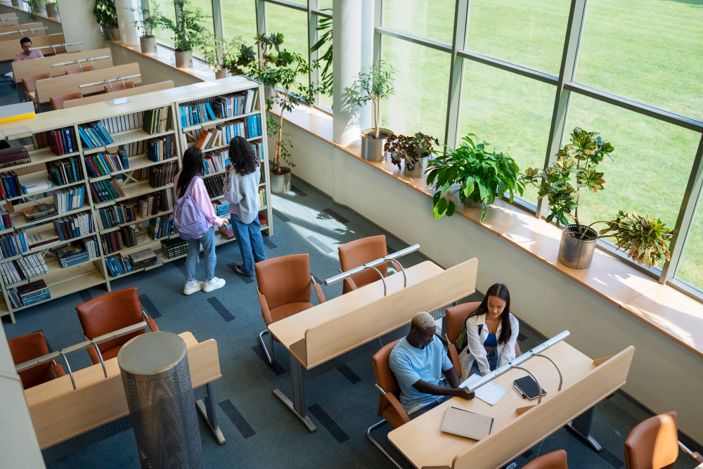 Vista aérea de uma biblioteca moderna e espaçosa com grandes janelas, onde estudantes estão sentados em mesas de estudo e outros estão olhando livros nas estantes.